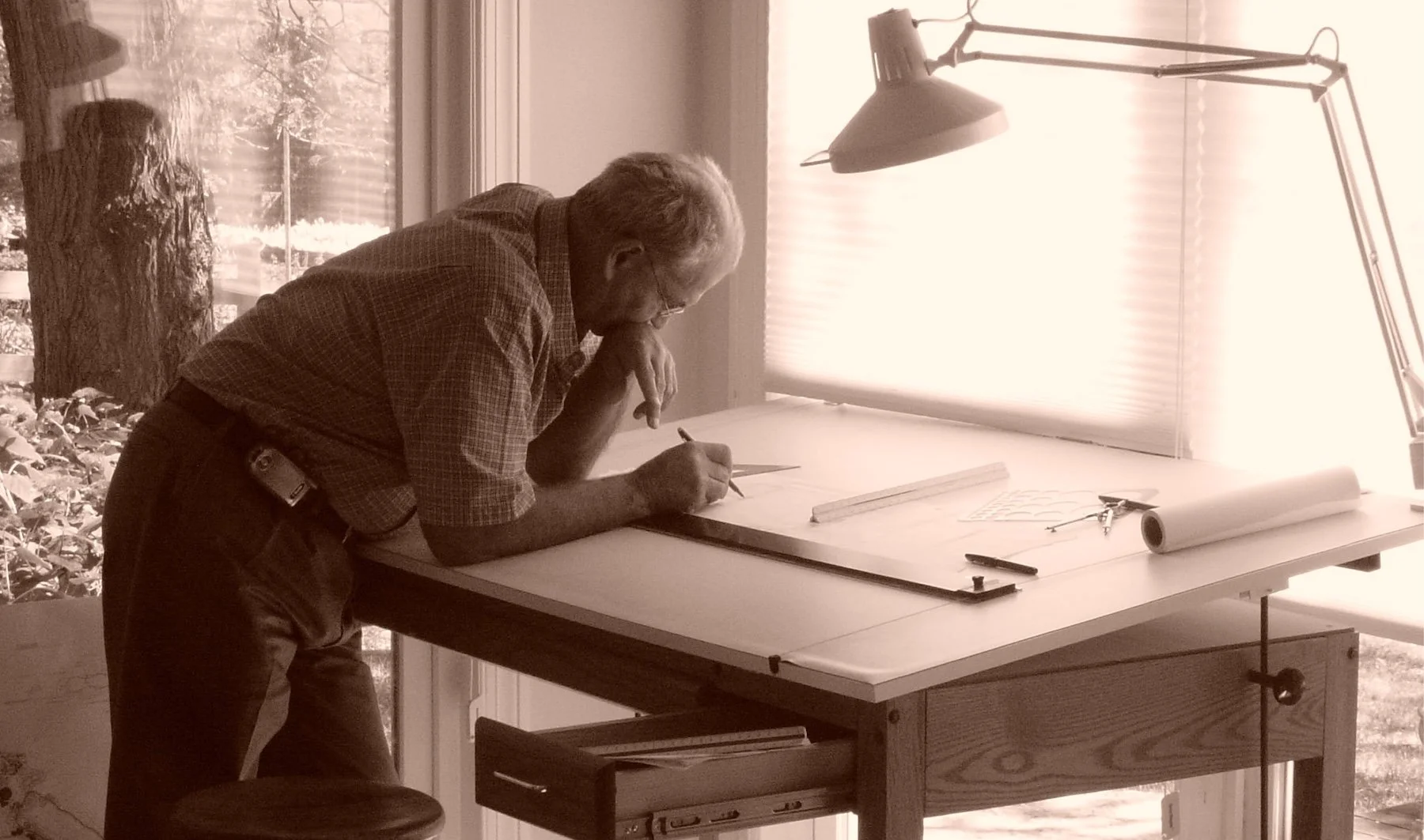 An older man with glasses and light-colored hair, wearing a checked shirt, is leaning over a drafting table, working on a drawing with a pencil