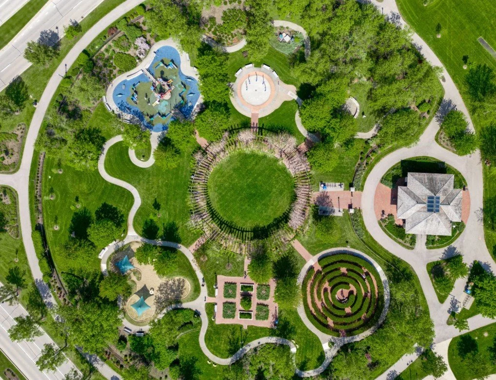 An aerial view of a park with various landscaped gardens, walkways, a playground with water features, a circular maze, and a building at the bottom right corner.