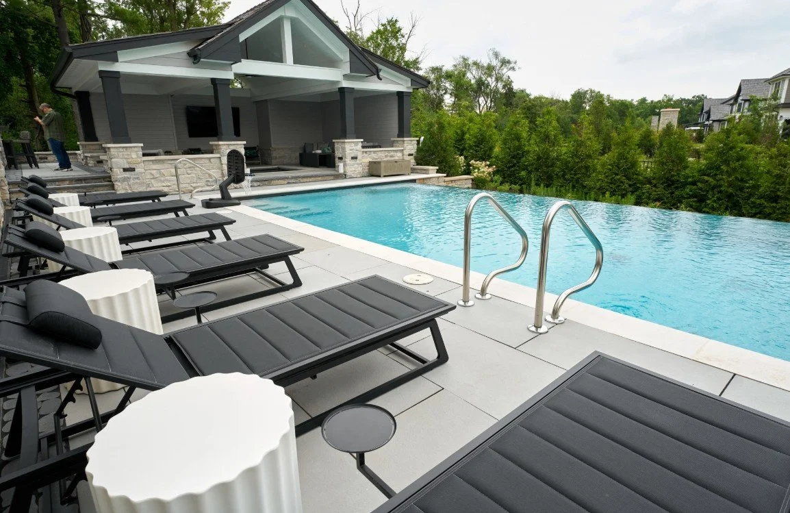 A modern backyard with a swimming pool, multiple black lounge chairs with pillows, and white side tables. In the background, there is a covered patio area with outdoor furniture and a stone exterior, surrounded by green trees and neighboring houses.