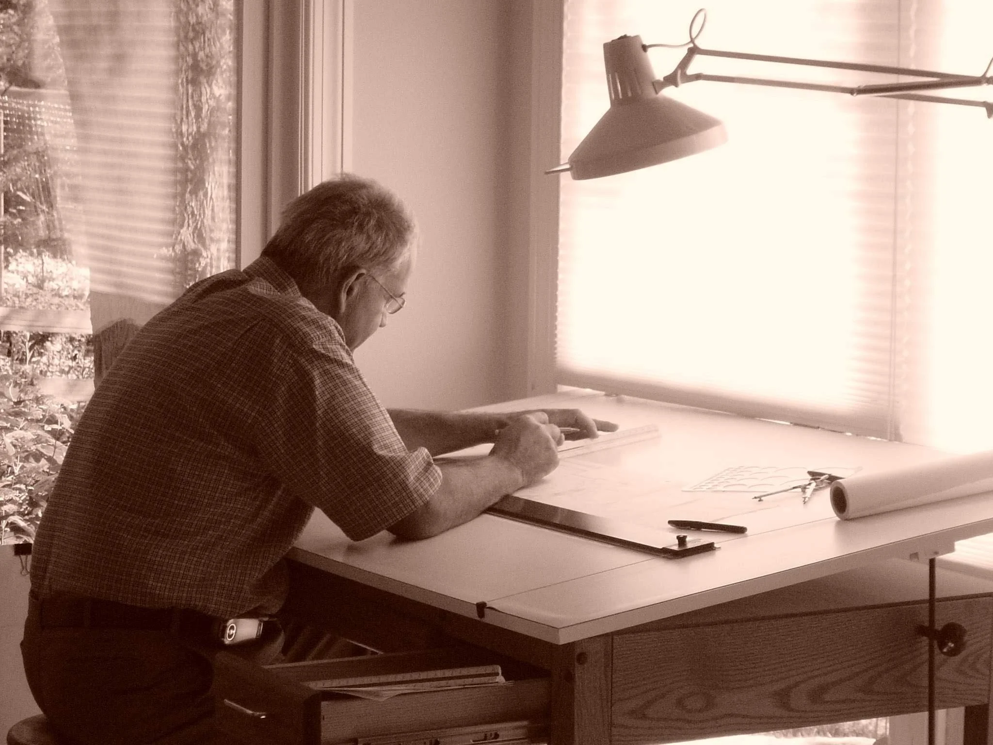 An elderly man with gray hair, glasses, and a checkered shirt sitting at a desk near a window, working on a drawing or writing with drawing tools and papers on the desk. A desk lamp illuminates the workspace.