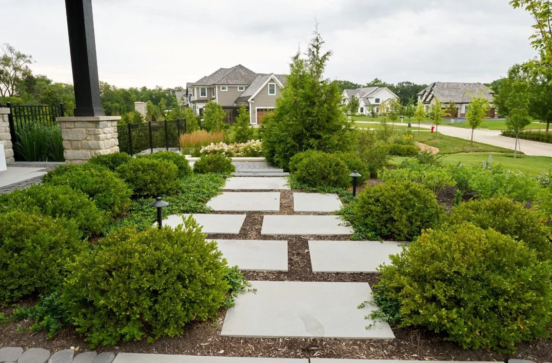 Residential front yard with a stone walkway, green bushes, small trees, and surrounding houses.