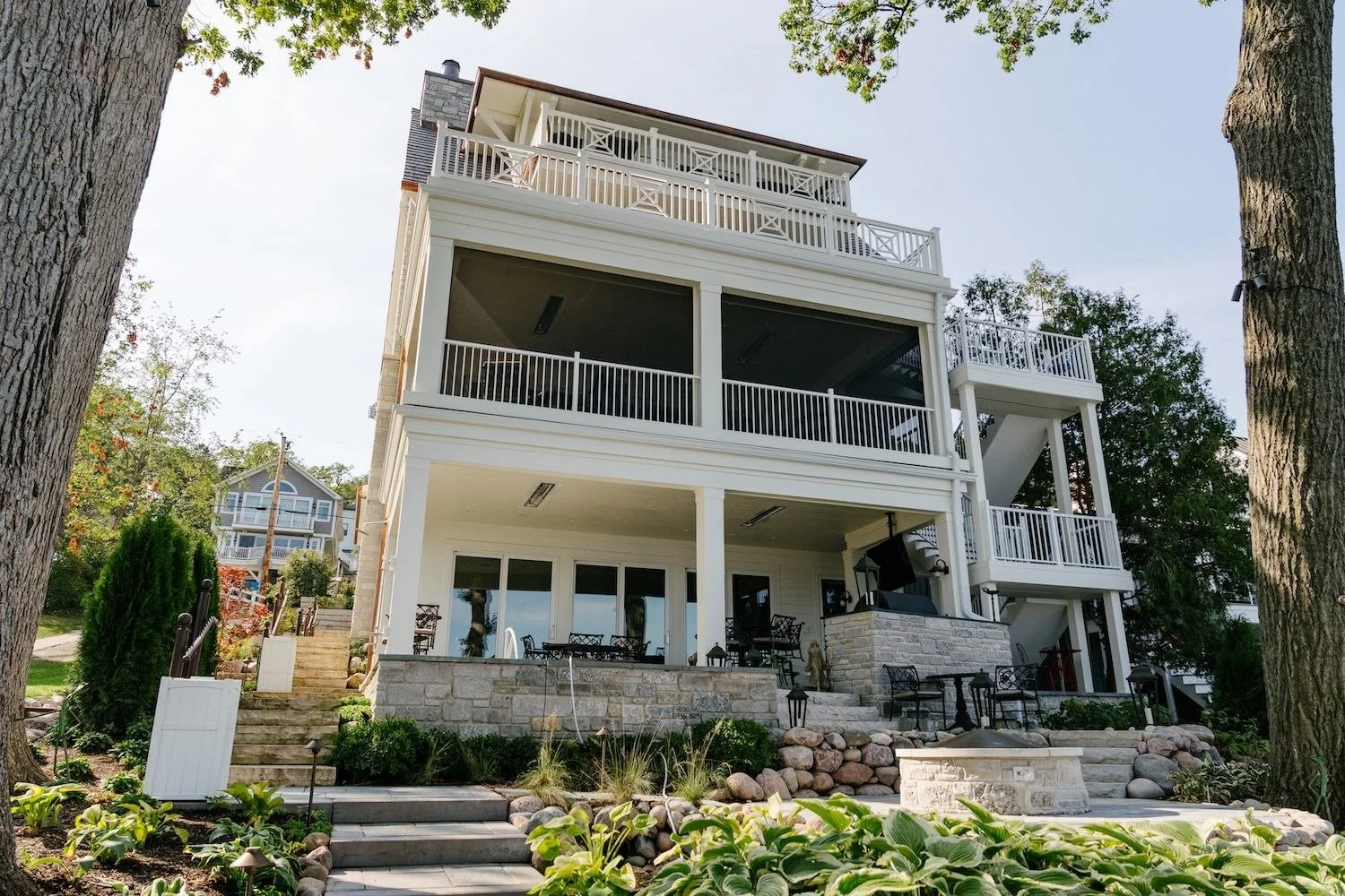Multi-story house with white balconies and outdoor seating area, surrounded by trees and landscaped garden with rocks and plants.