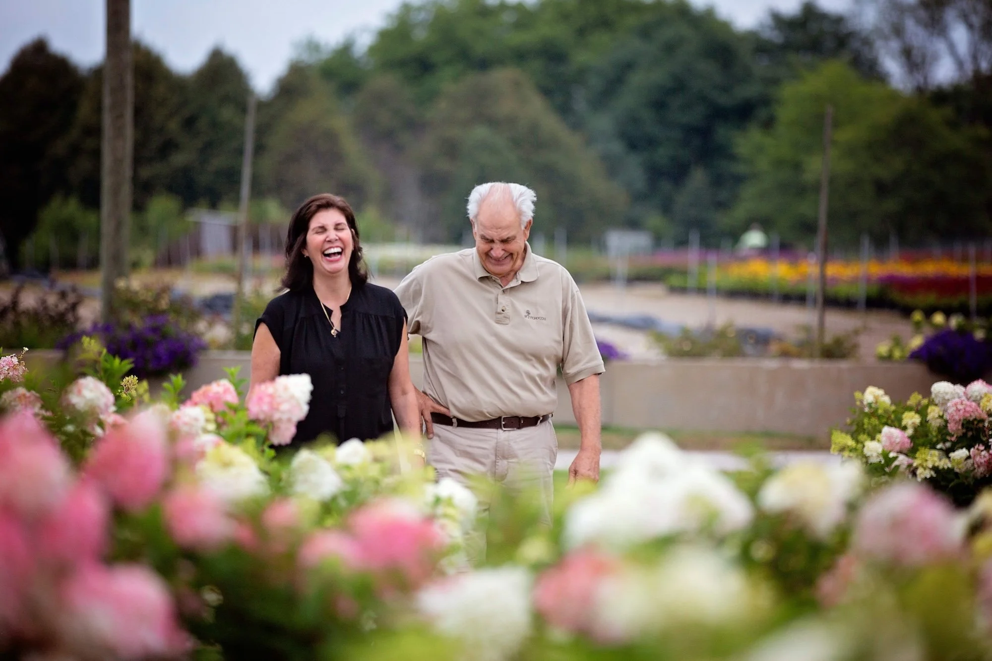 A smiling woman and man walking together among colorful flowers outdoors in a garden or park, with trees and pathways in the background.