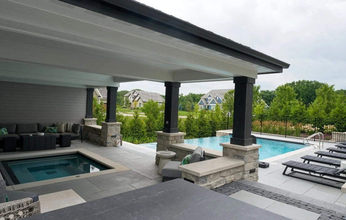 Outdoor swimming pool area with lounge chairs, a hot tub, and shaded seating under a covered patio, surrounded by greenery and neighboring houses in the distance.
