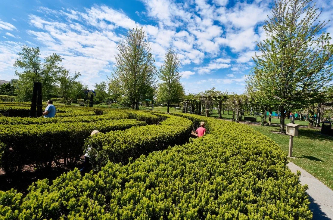 People walking through a well-maintained garden with green bushes, trees, and a blue sky with scattered clouds.
