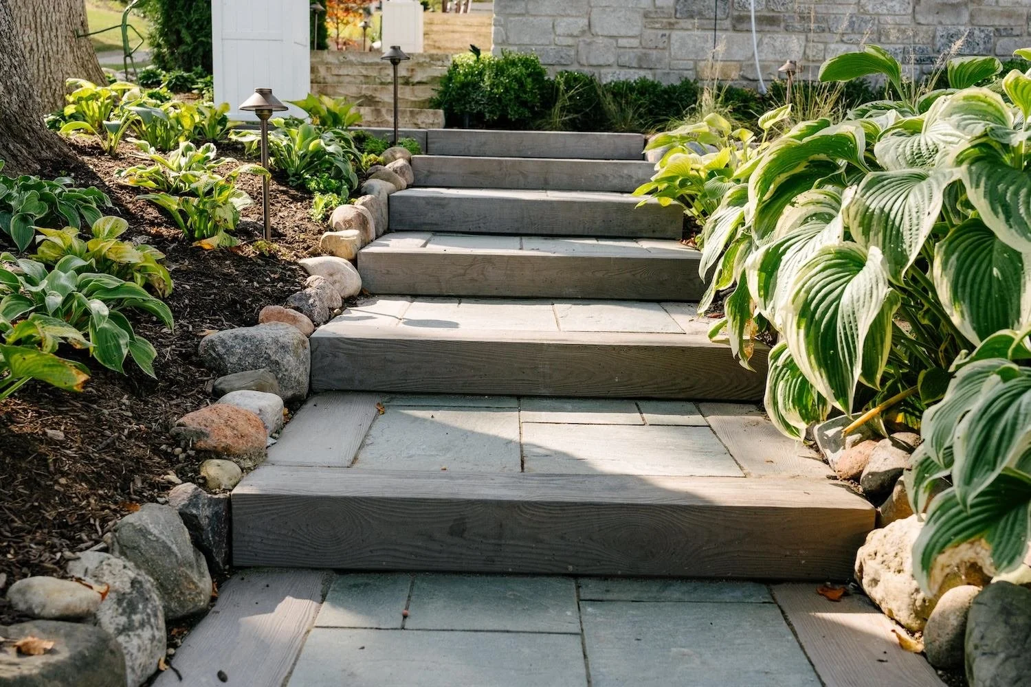 Wooden steps leading up to a house entrance, flanked by green plants and small rocks, with pathway lights on either side of the stairs.