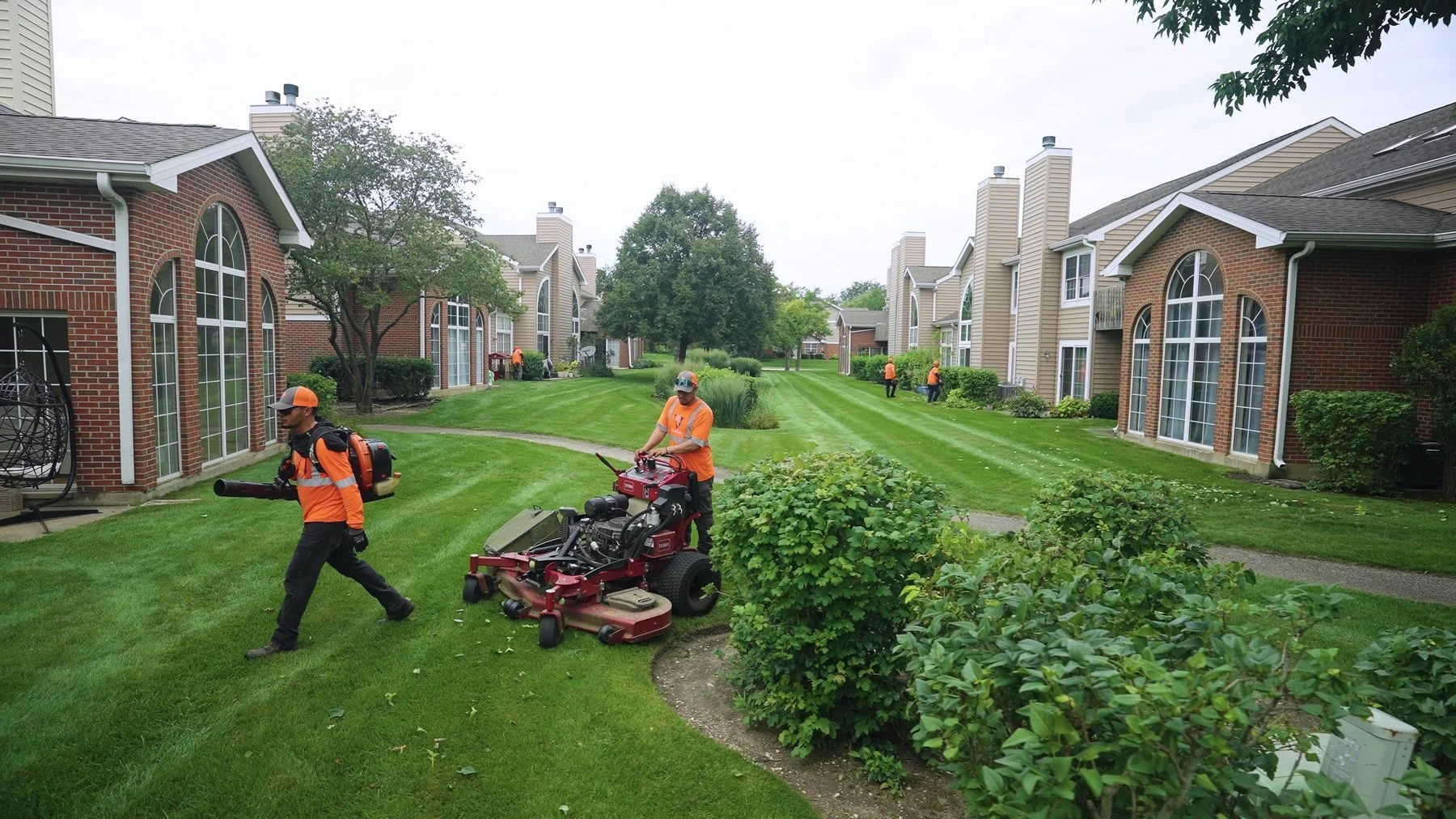 People mowing and maintaining a well-manicured lawn in a residential community with brick and siding houses, some with large arched windows, and trees lining the street.