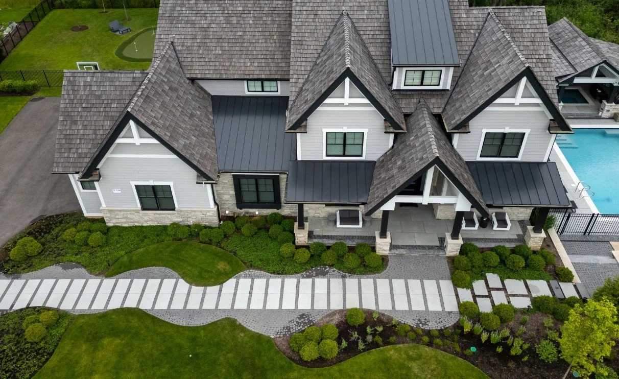 Aerial view of a modern house with a grey roof, white siding, and a backyard swimming pool, surrounded by a well-maintained lawn and garden.