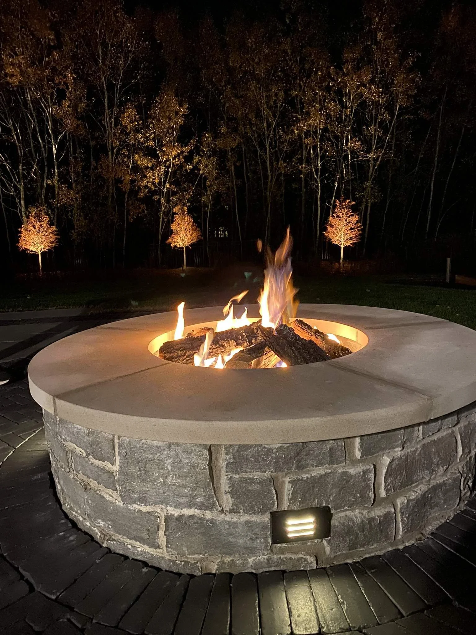 Nighttime scene featuring a stone fire pit with a controlled fire burning inside. In the background, there are trees with autumn foliage illuminated by outdoor lighting.