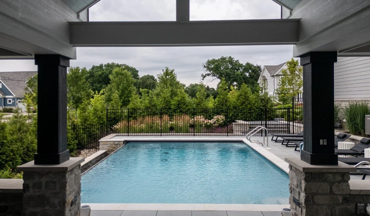 A backyard pool area under a covered patio, with lounge chairs and a black metal fence, surrounded by trees and residential houses.