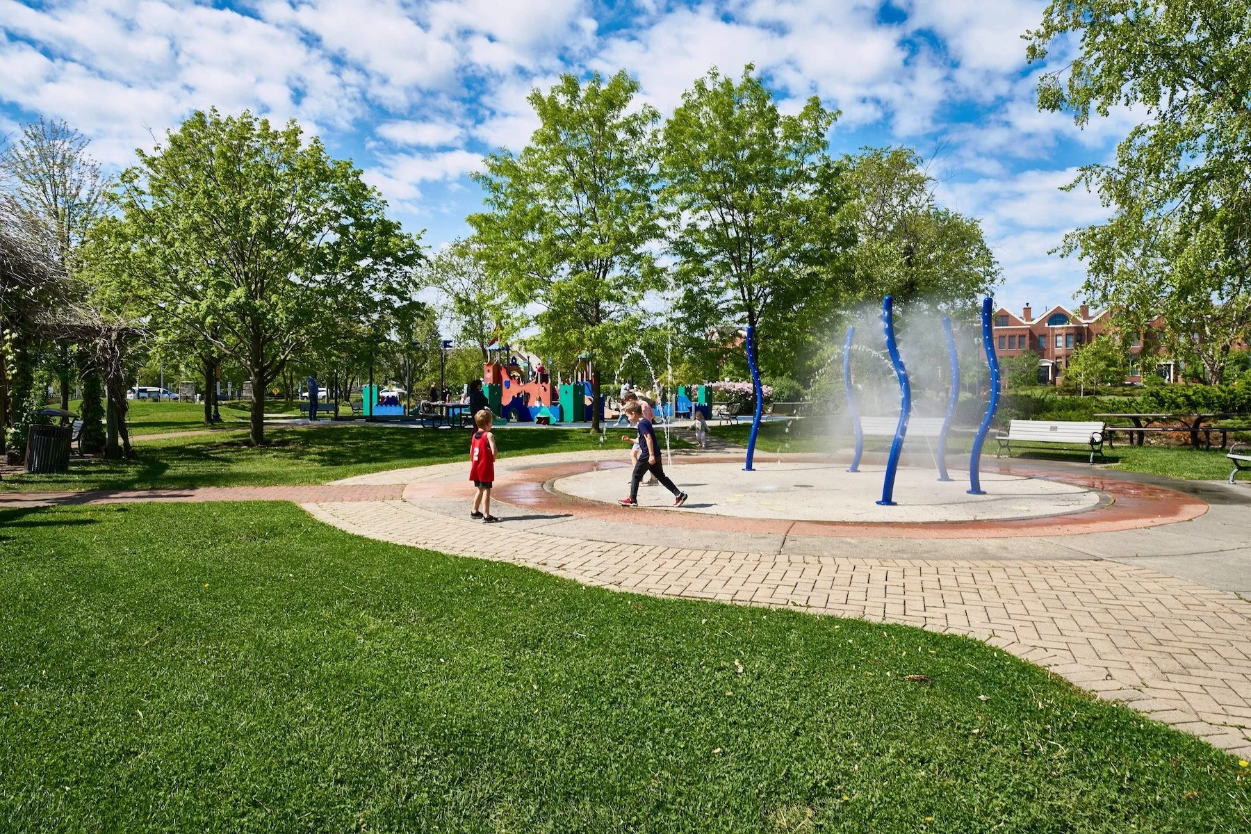 Children playing at a splash pad in a park with trees, benches, and a playground in the background.