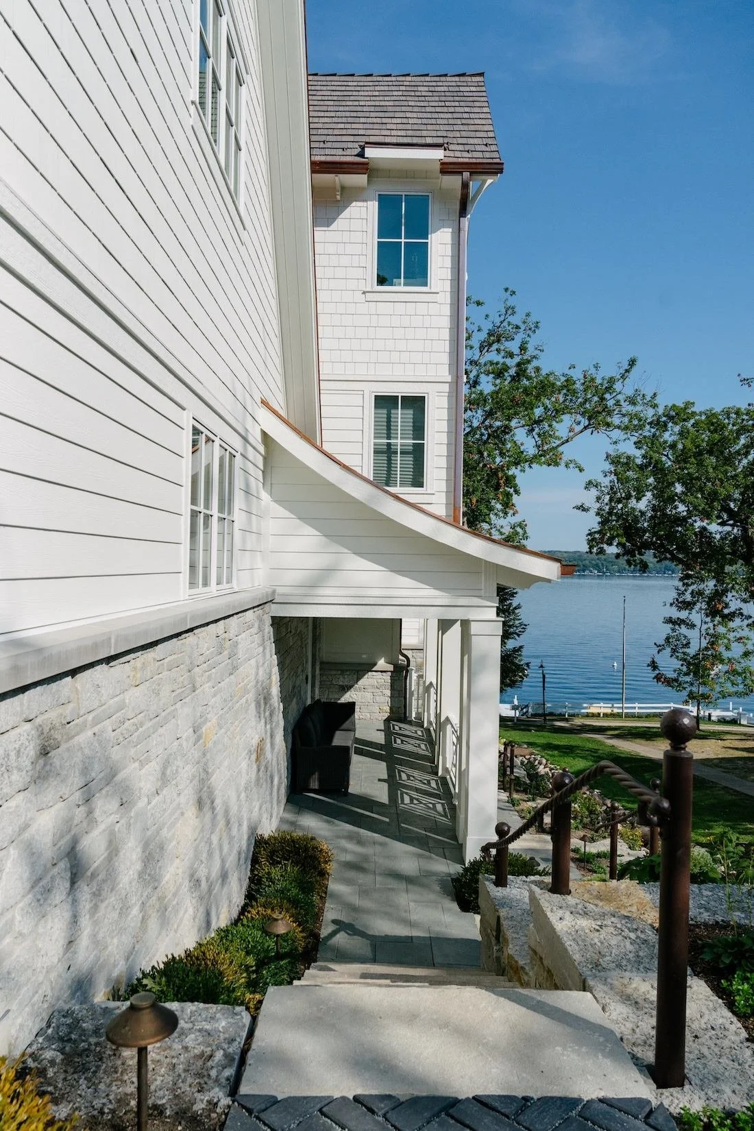 Side view of a white house with multiple windows overlooking a body of water, with a small patio area and steps leading down to a green lawn and waterfront.