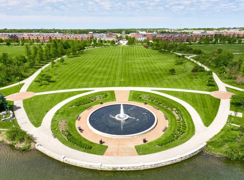 Aerial view of a park with a large grassy field, walking paths, trees, a circular fountain, and benches, with a residential area in the background.