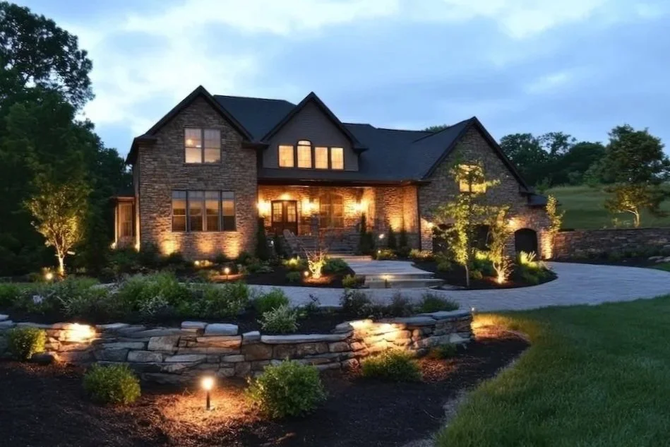 A two-story brick house with multiple windows, illuminated from inside, surrounded by landscaped garden with trees, shrubs, and pathway lighting at dusk.