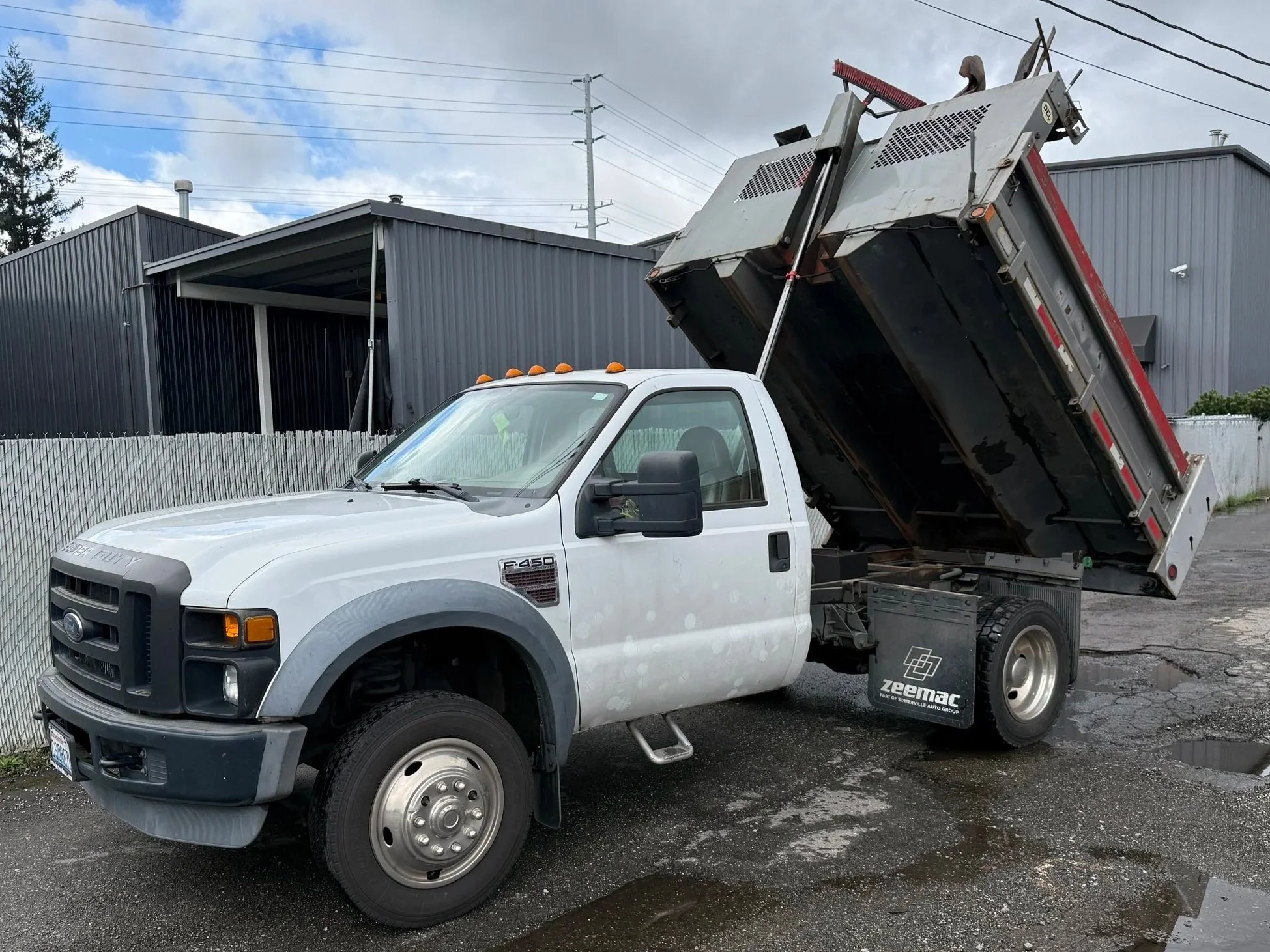 A white dump truck with the bed raised, in the middle of fixing potholes on an asphalt parking lot.