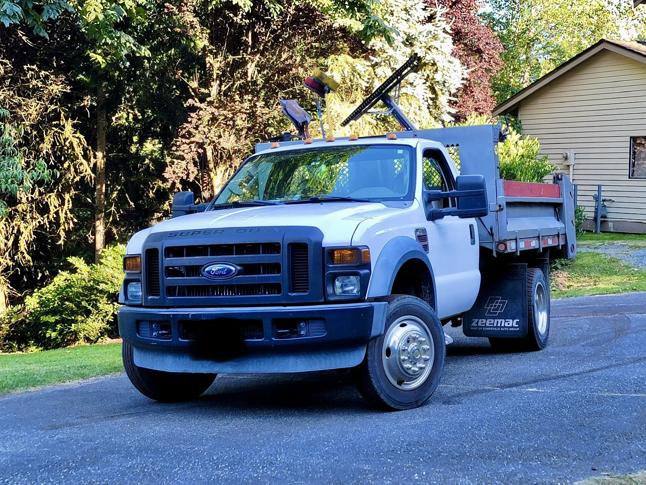A white dump truck parked on a paved driveway with asphalt rakes and shovels sticking up.