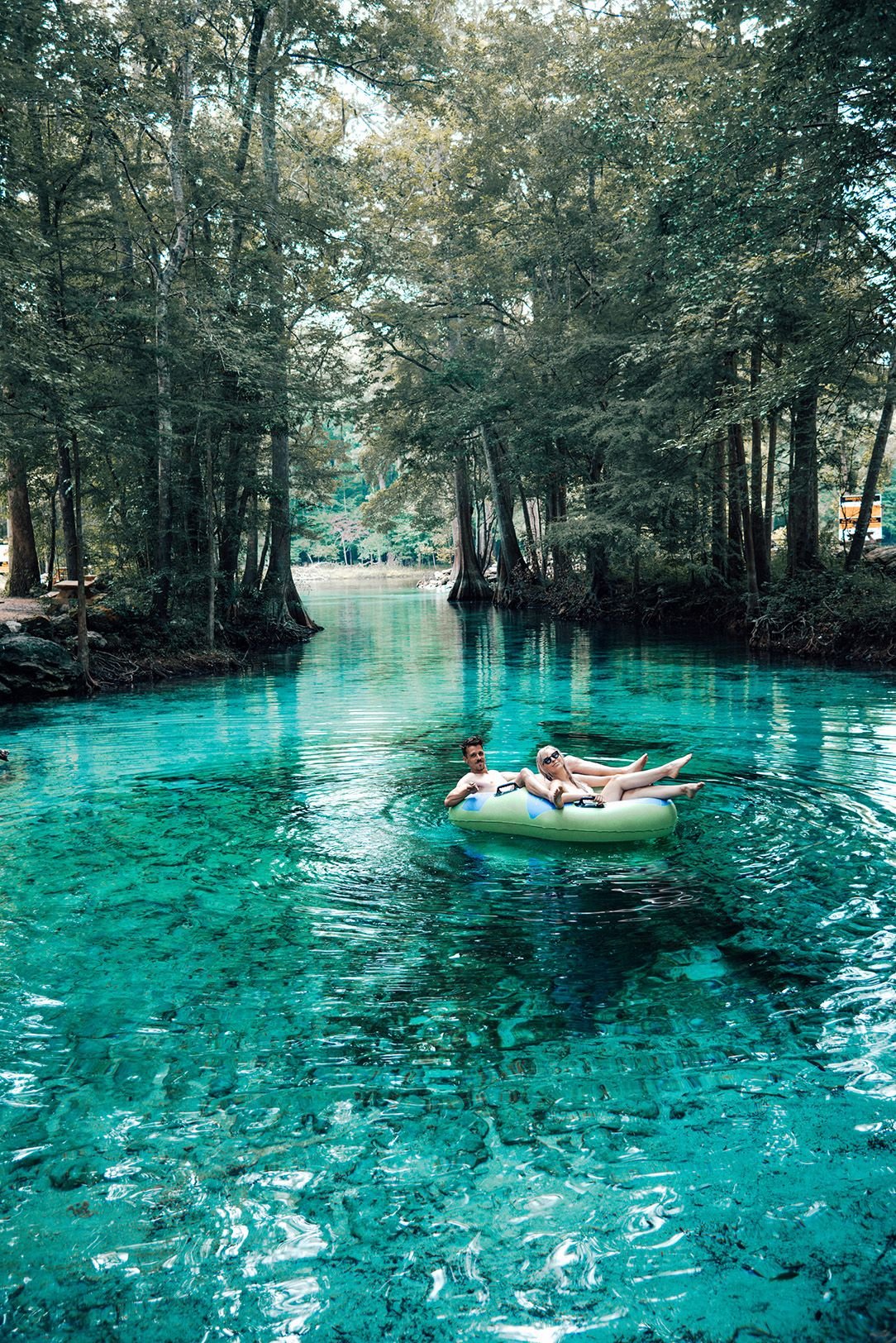 Two people in swimsuits relaxing on an inflatable raft in a clear, blue-green river surrounded by dense trees in a forest setting.