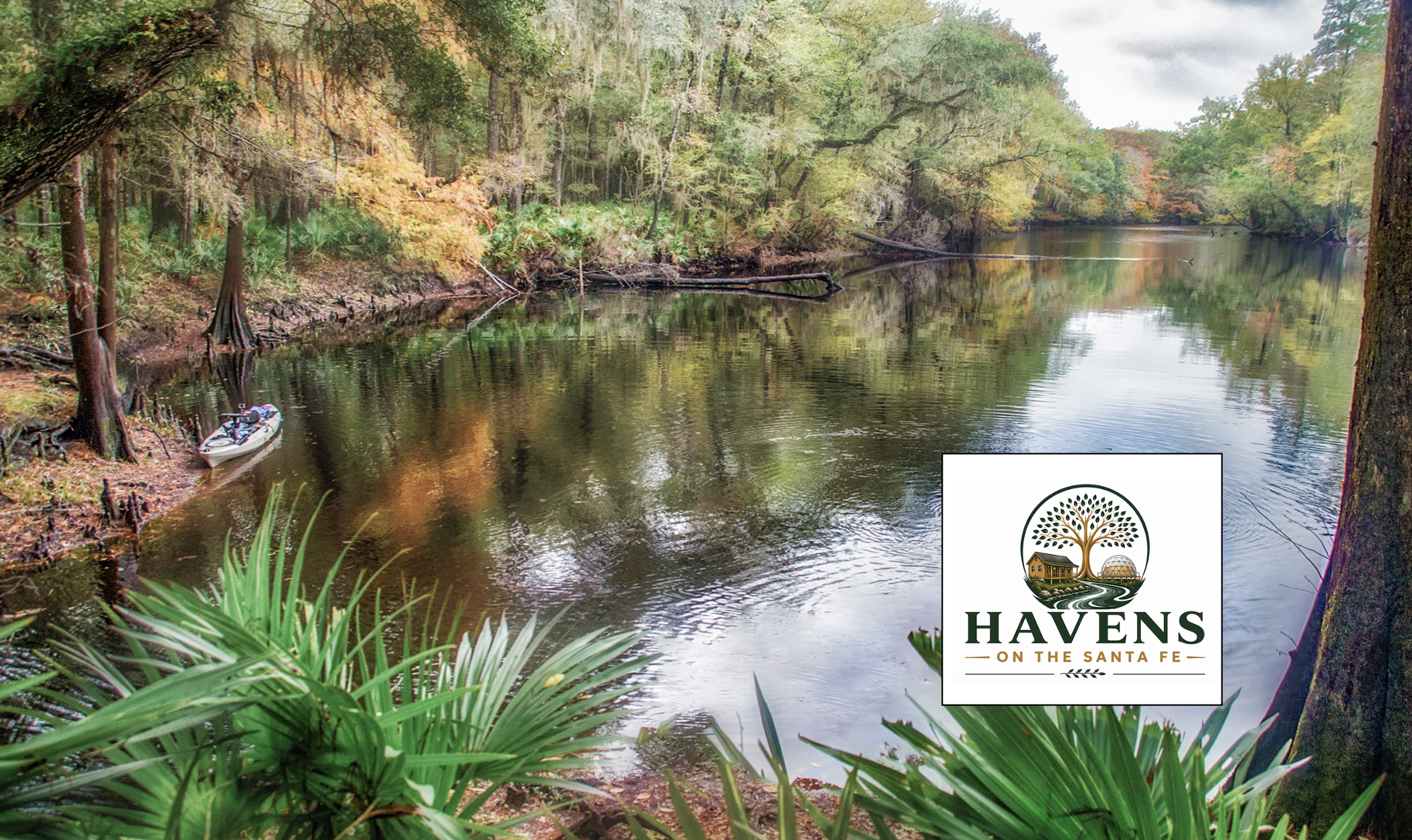 A river flowing through a lush green forest with trees on both sides and a small boat docked on the muddy riverbank. The scene is serene and natural, with reflections of the trees in the water. There is a logo in the bottom right corner that reads 'Havens on the Santa Fe' with a tree, a house, and a globe.