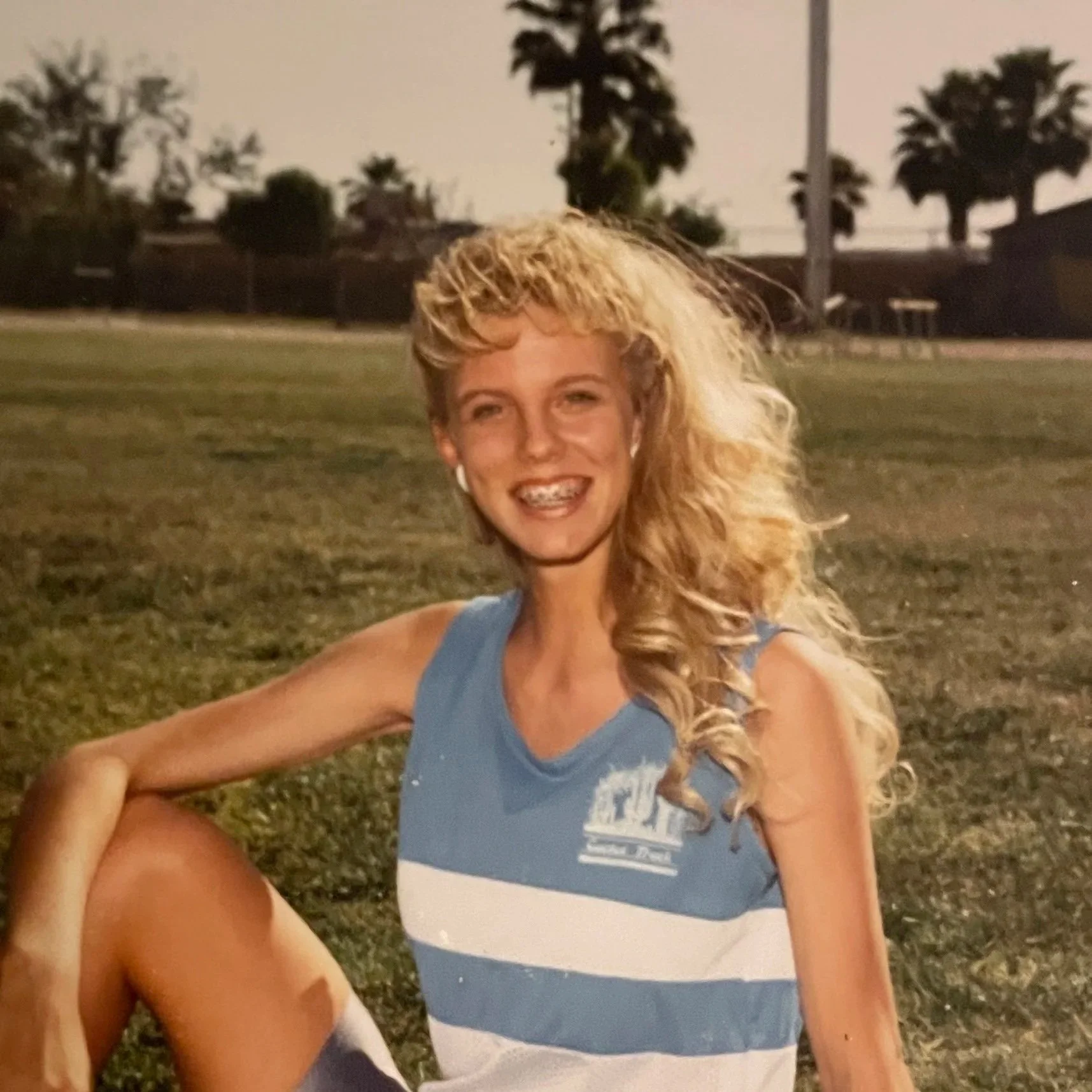 A woman with long, curly blonde hair wearing a blue sleeveless top, smiling with braces, sitting outdoors on grass during daytime with trees in the background.