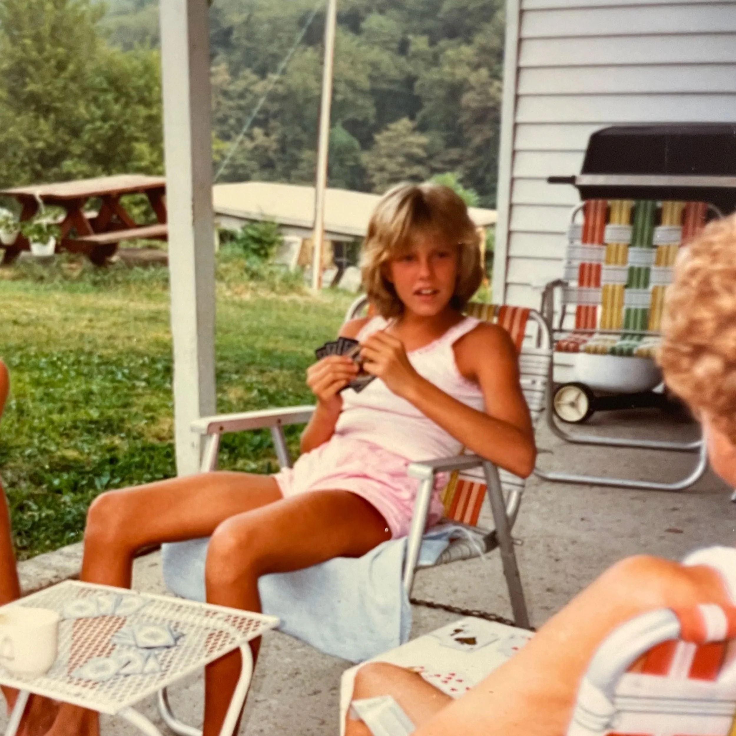 A young girl with shoulder-length blonde hair sitting on a striped lawn chair on a porch, holding playing cards, with outdoor furniture and greenery visible in the background.