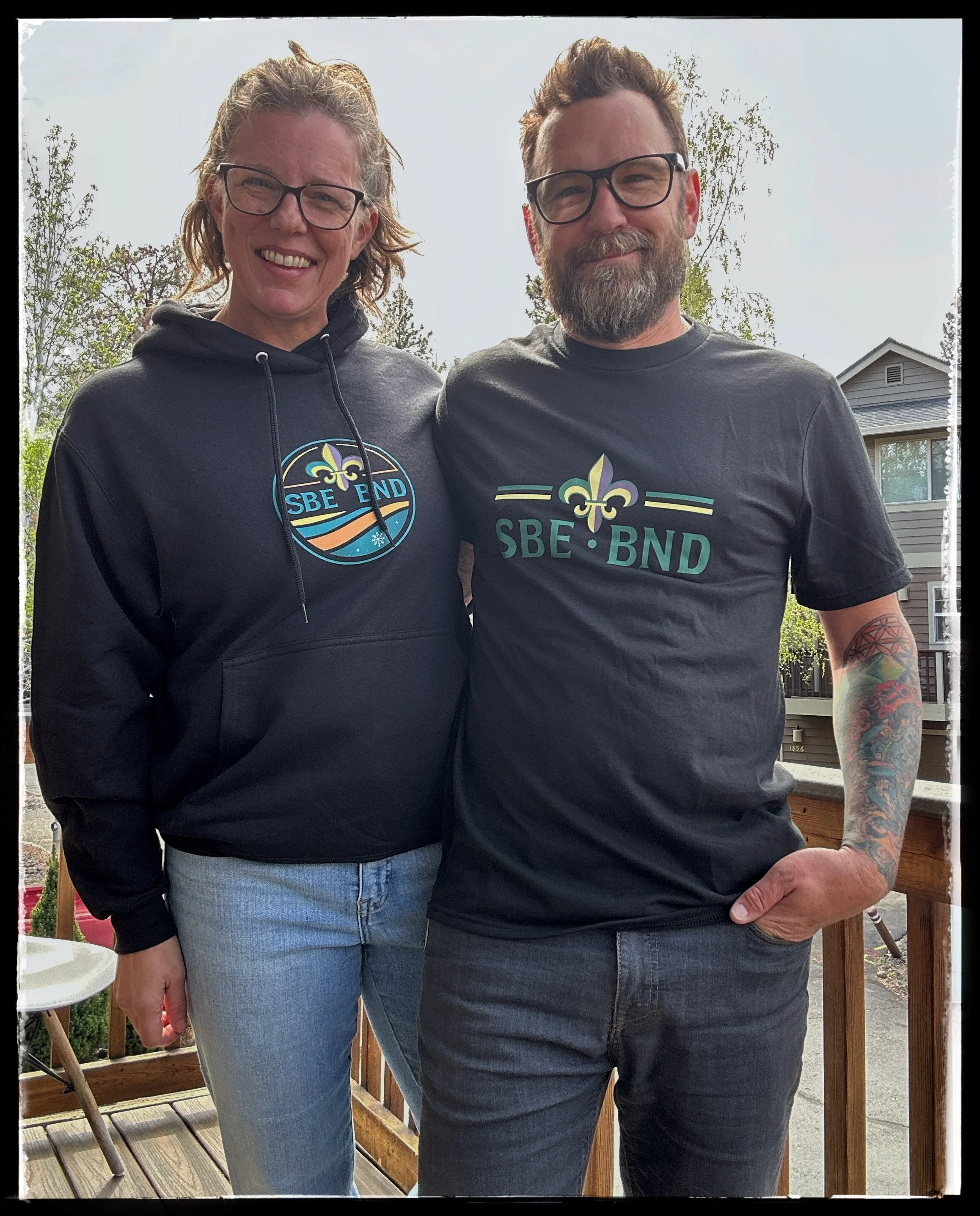 A woman and a man standing close together on a wooden deck, smiling at the camera. The woman is wearing a black hoodie with a logo, light blue jeans, and glasses. The man has a beard, glasses, a black t-shirt with a logo, and tattoos on his right arm.