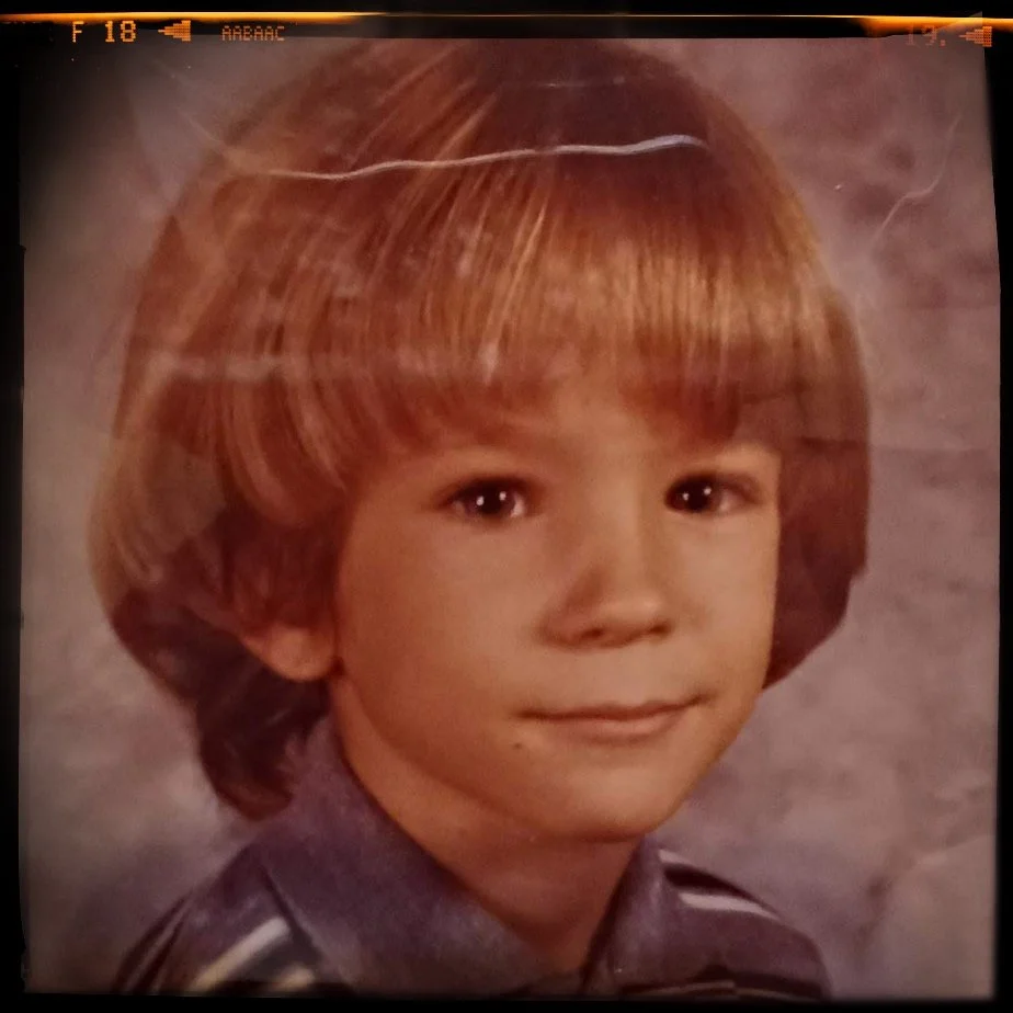 School portrait of a young boy with light brown, shoulder-length hair and a slight smile, wearing a gray shirt with a collar.