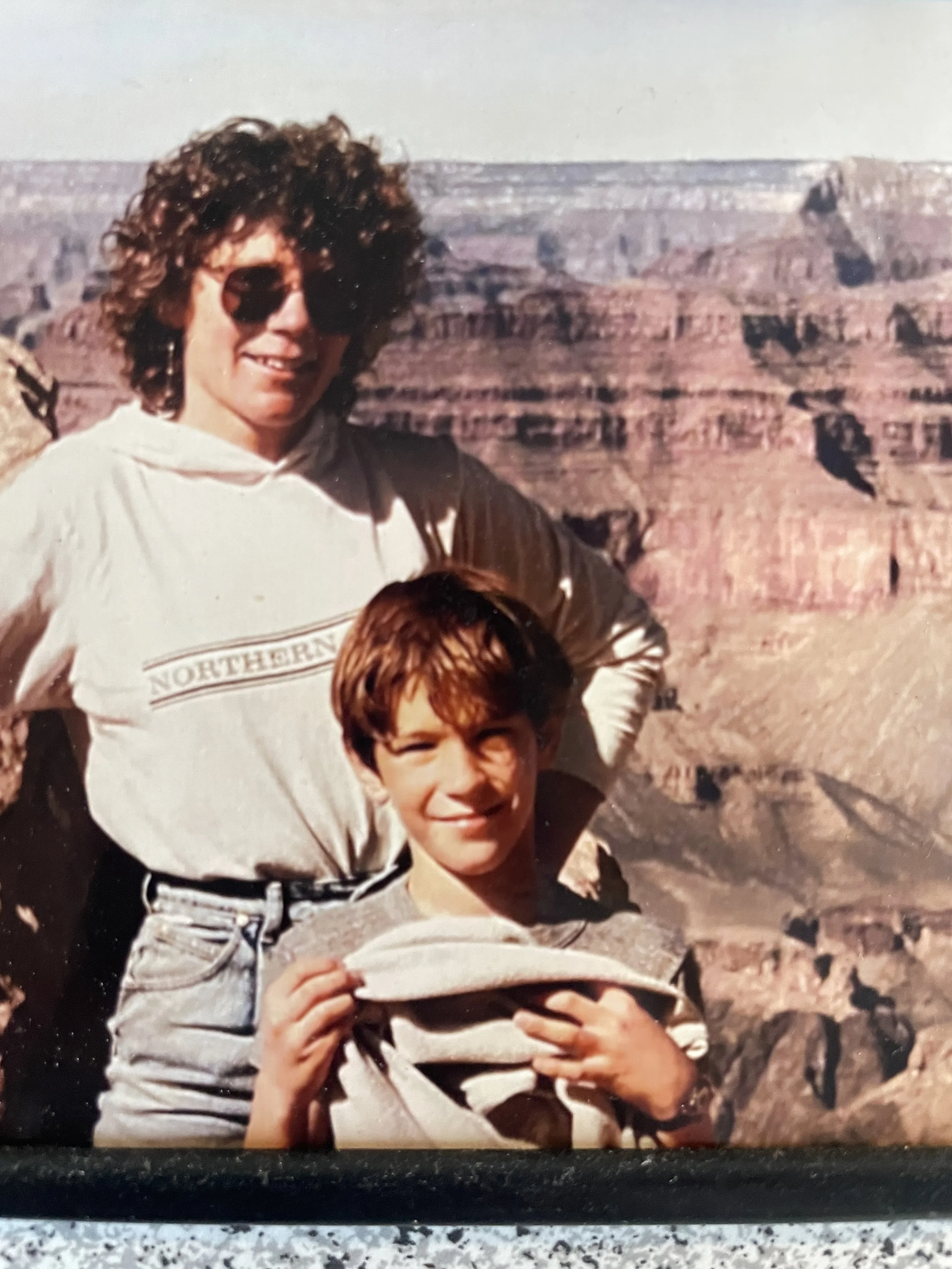 A woman with curly hair wearing sunglasses and a white top standing outdoors near the Grand Canyon, holding a piece of paper or photograph, with a child in front of her.