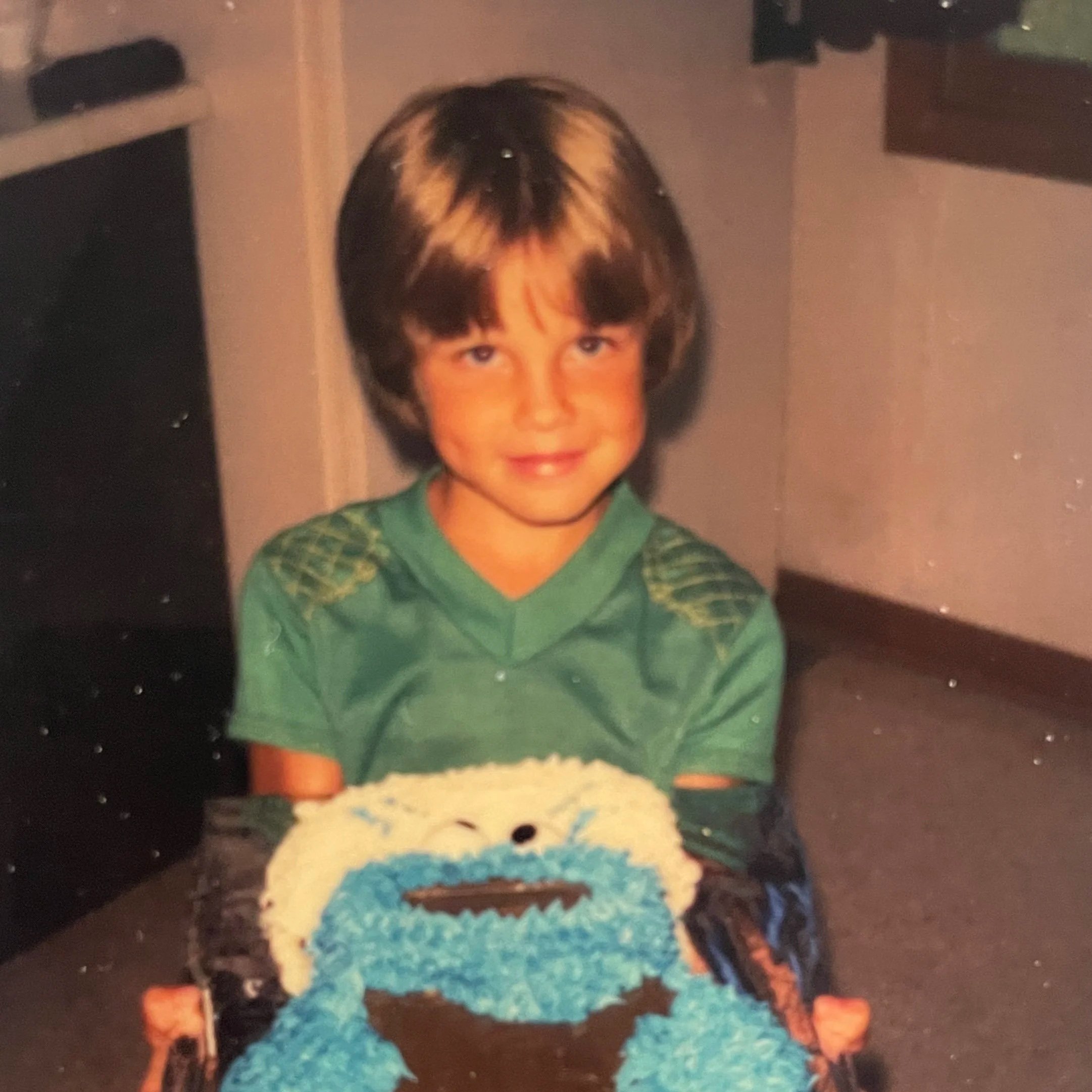 Child with short brown hair and blue eyes wearing a green shirt, standing indoors near a blue ball and colorful toy blocks.