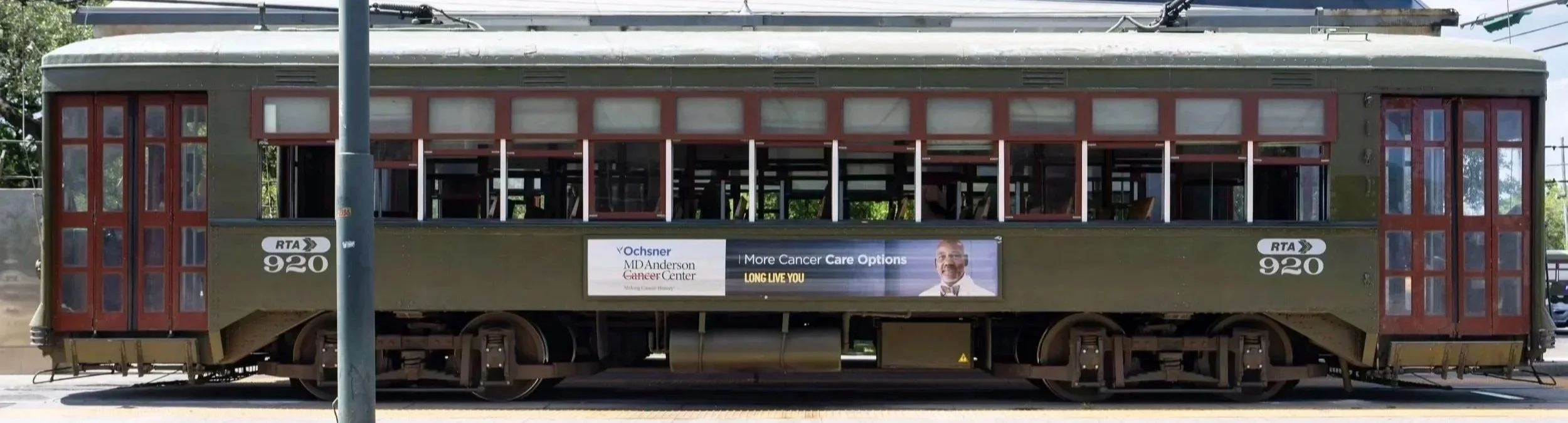 An old-fashioned trolley with a green and red exterior, featuring large windows and a sign with advertisements for a cancer center.