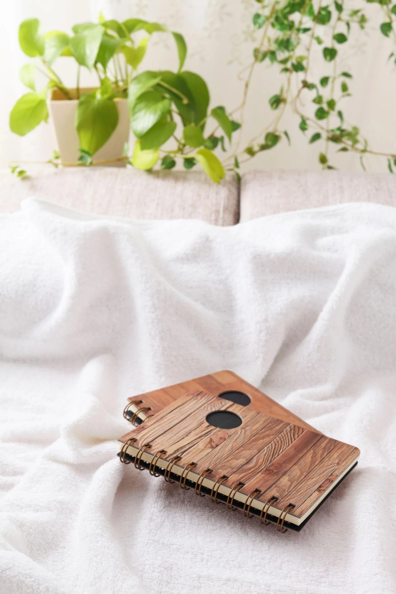 A wooden spiral-bound notebook with a woodgrain pattern on the cover, resting on a white textured fabric surface, with a background of green houseplants and a pinkish-beige sofa.