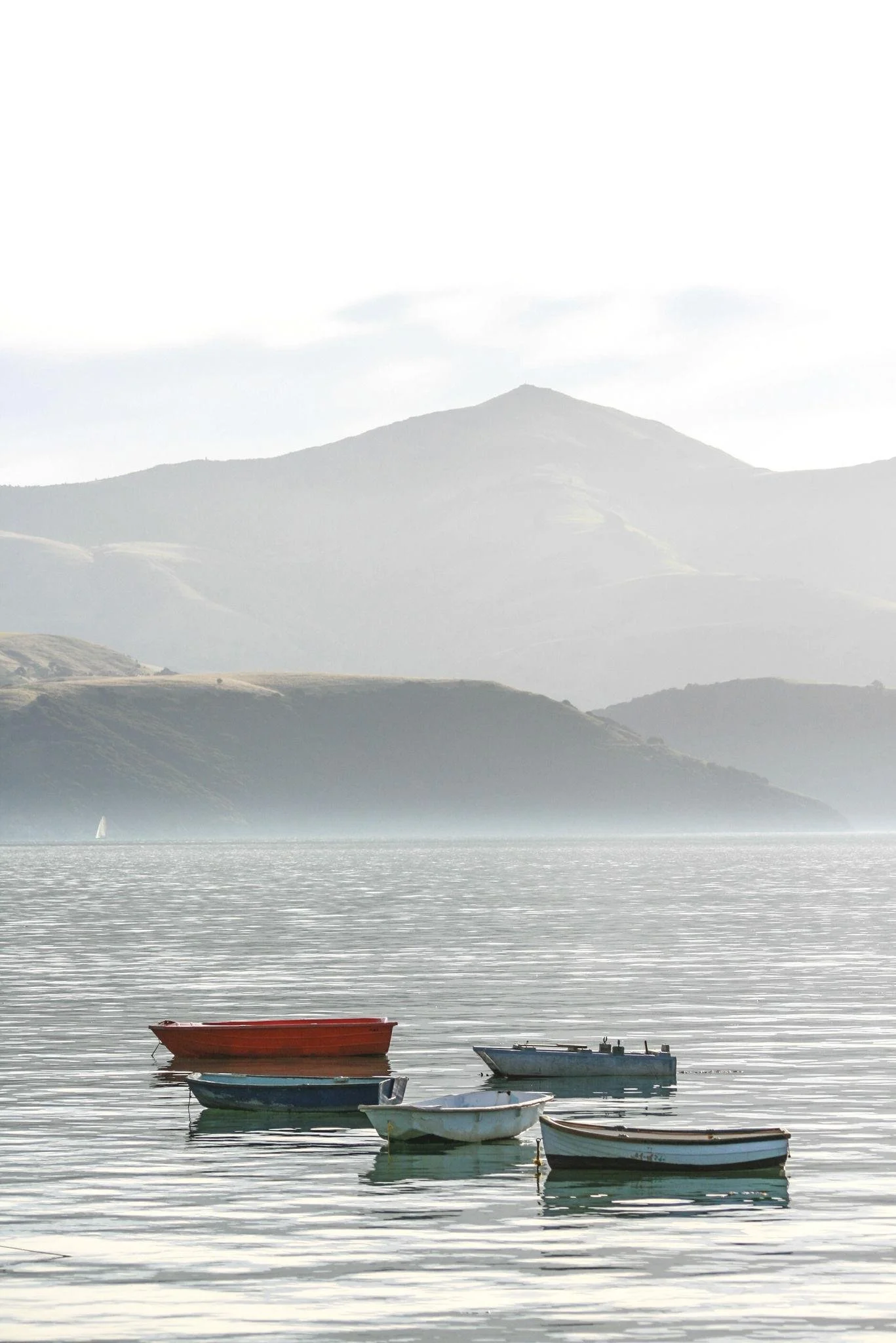 A calm body of water with four small boats floating near the shore, and mountains in the background under a cloudy sky.