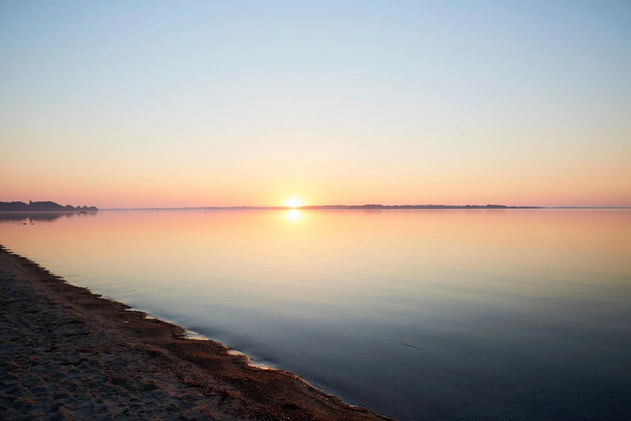 Sunset over calm river with sandy shore in foreground and trees in the distance.