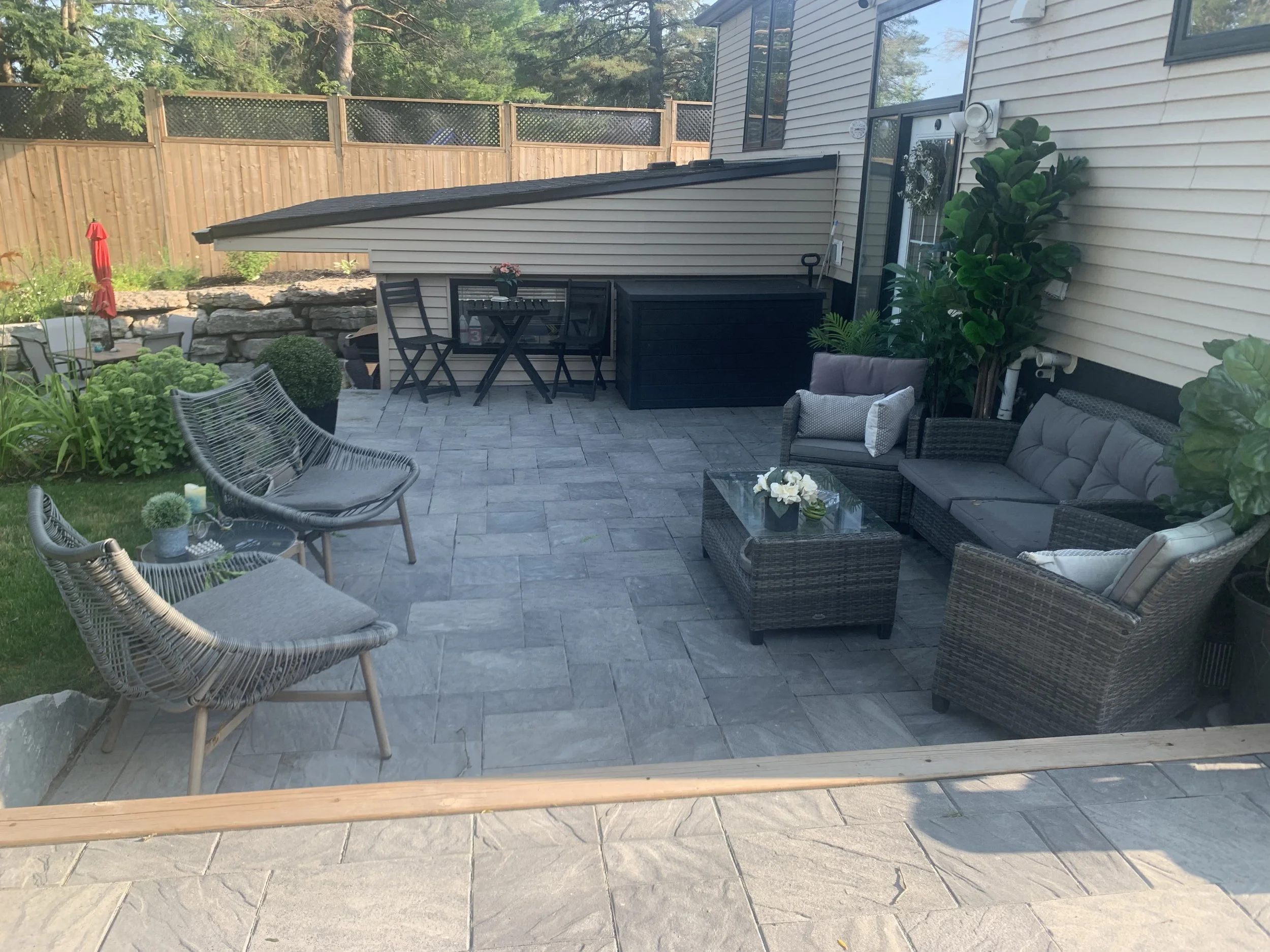 A backyard patio with outdoor furniture, a stone retaining wall, a wooden fence, lush green plants, and a table with an umbrella visible in the background.