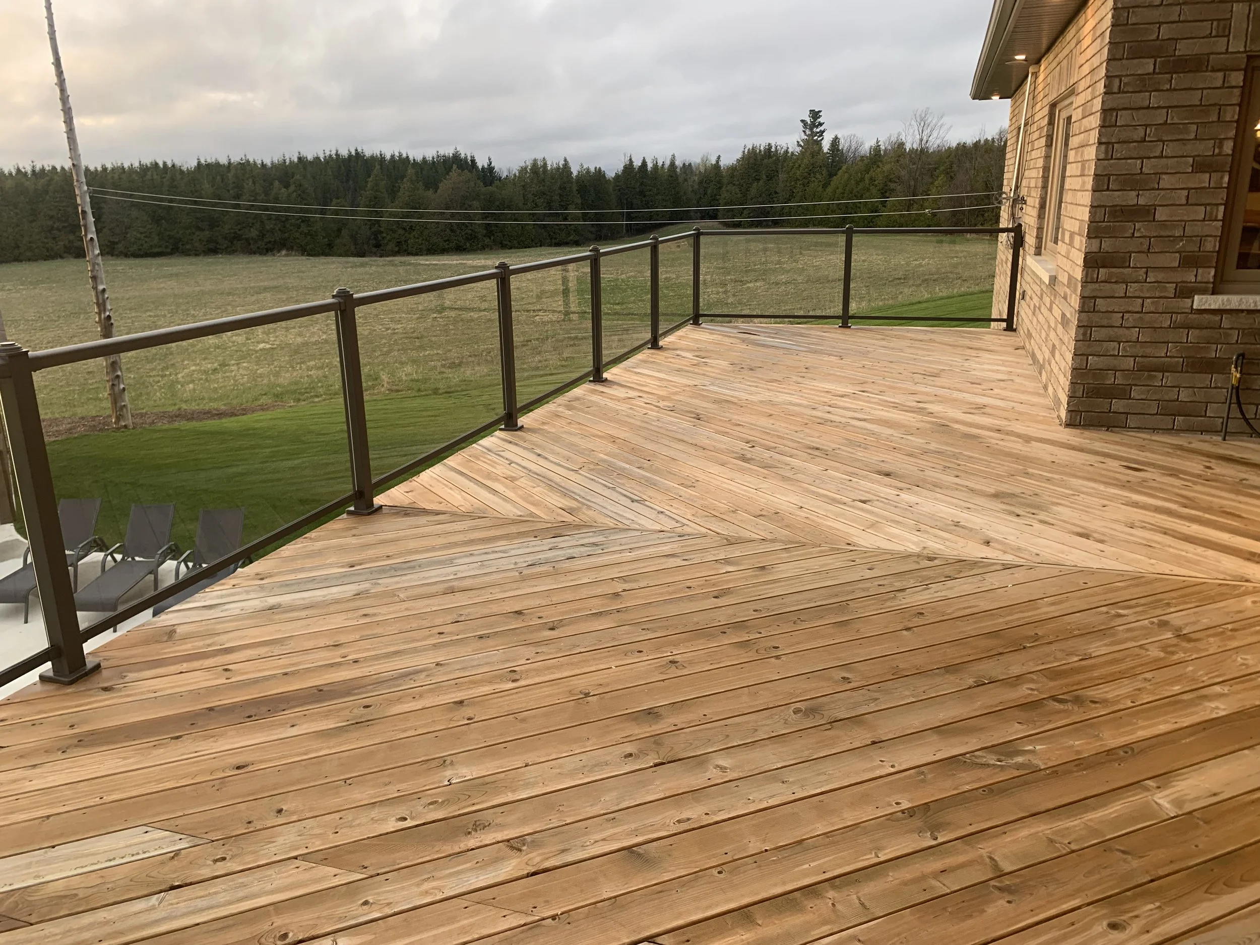 A wooden deck attached to a house with a railing, overlooking a grassy field with trees in the background and an overcast sky.