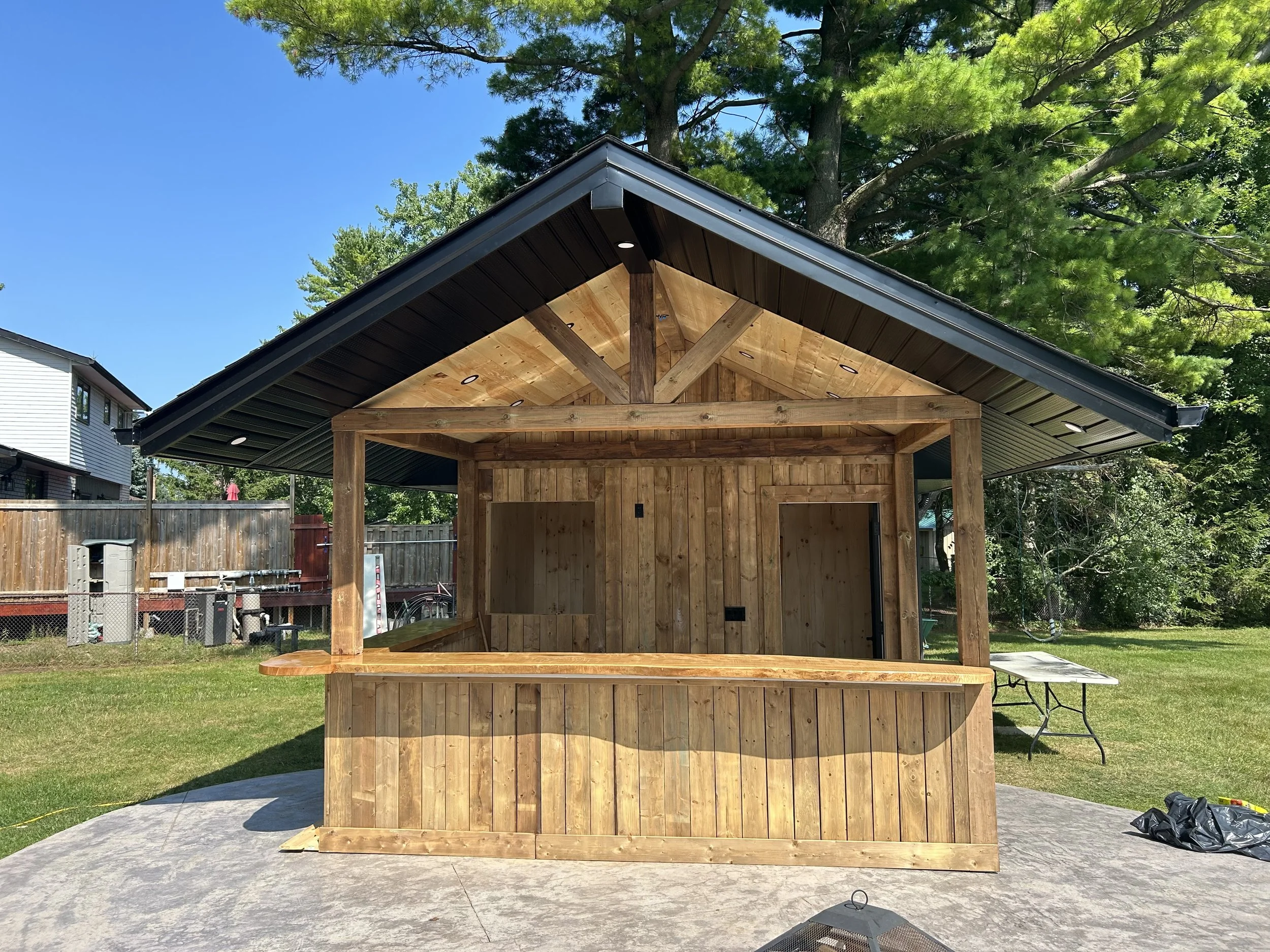 A small wooden gazebo with a black metal roof is under construction in a backyard. The structure has an open front, a wood-paneled ceiling, and wooden support posts. The backyard is grassy with neighboring houses and tall green trees in the backgroun