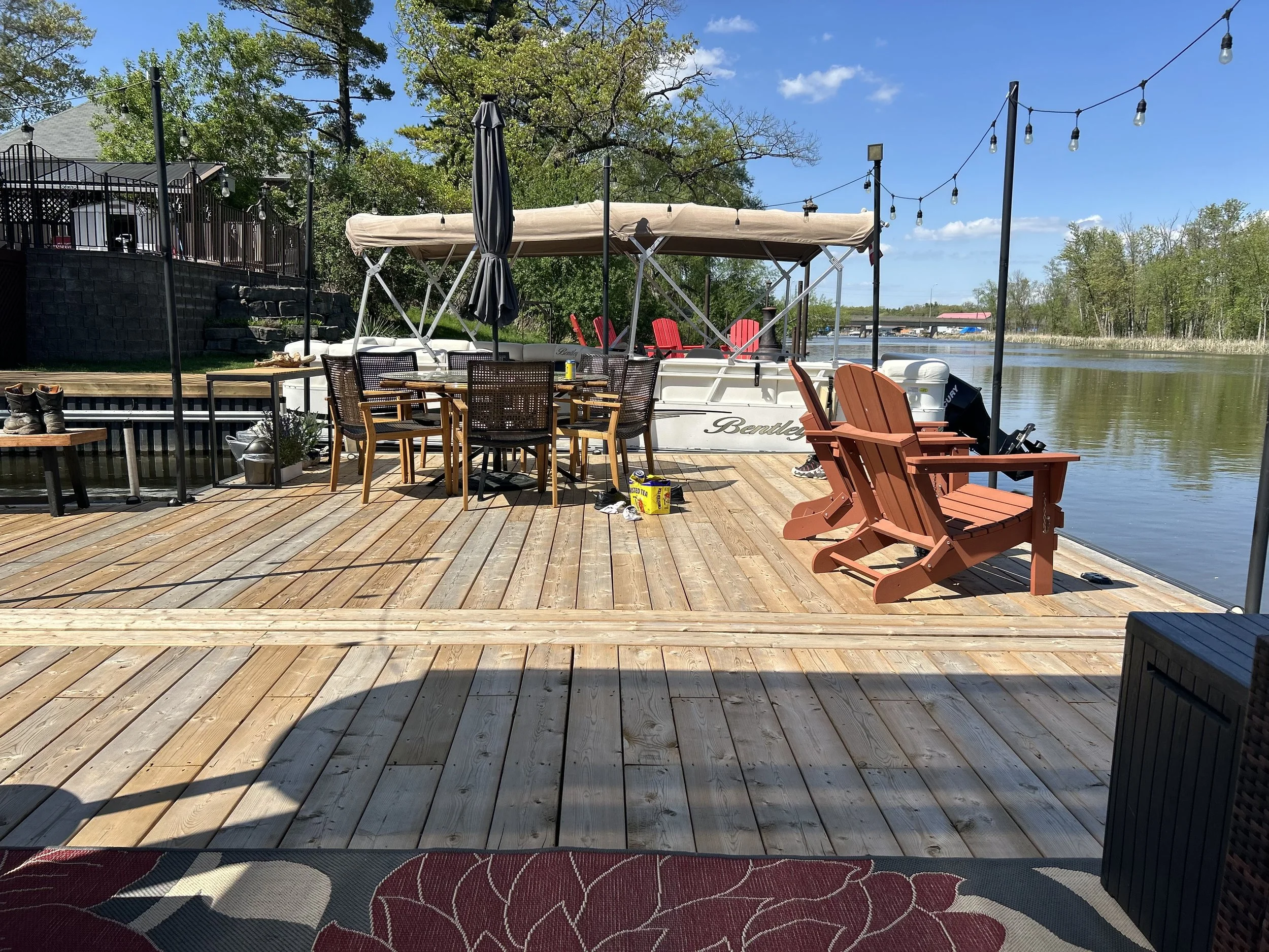A wooden dock by a calm river with outdoor furniture including a table with chairs, Adirondack chairs, a boat, and string lights overhead under a partly cloudy blue sky.