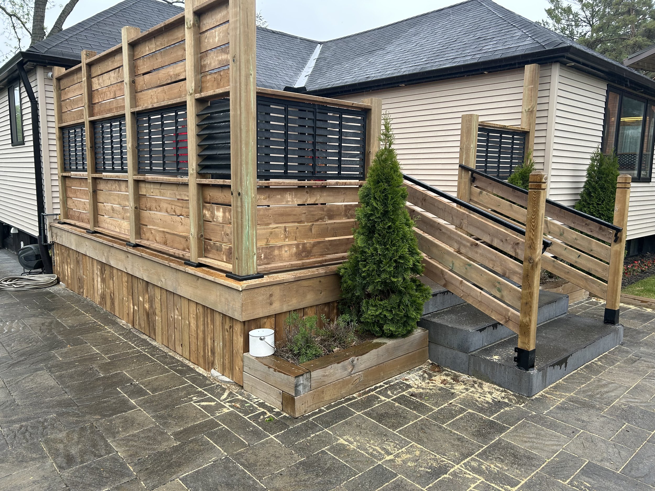 A house with beige siding and large windows, featuring a newly built wooden deck with fencing. The deck has a black privacy screen, a ramp, and is elevated on concrete blocks. There are small evergreen shrubs planted around the deck, and a stone-paved patio area in the foreground.