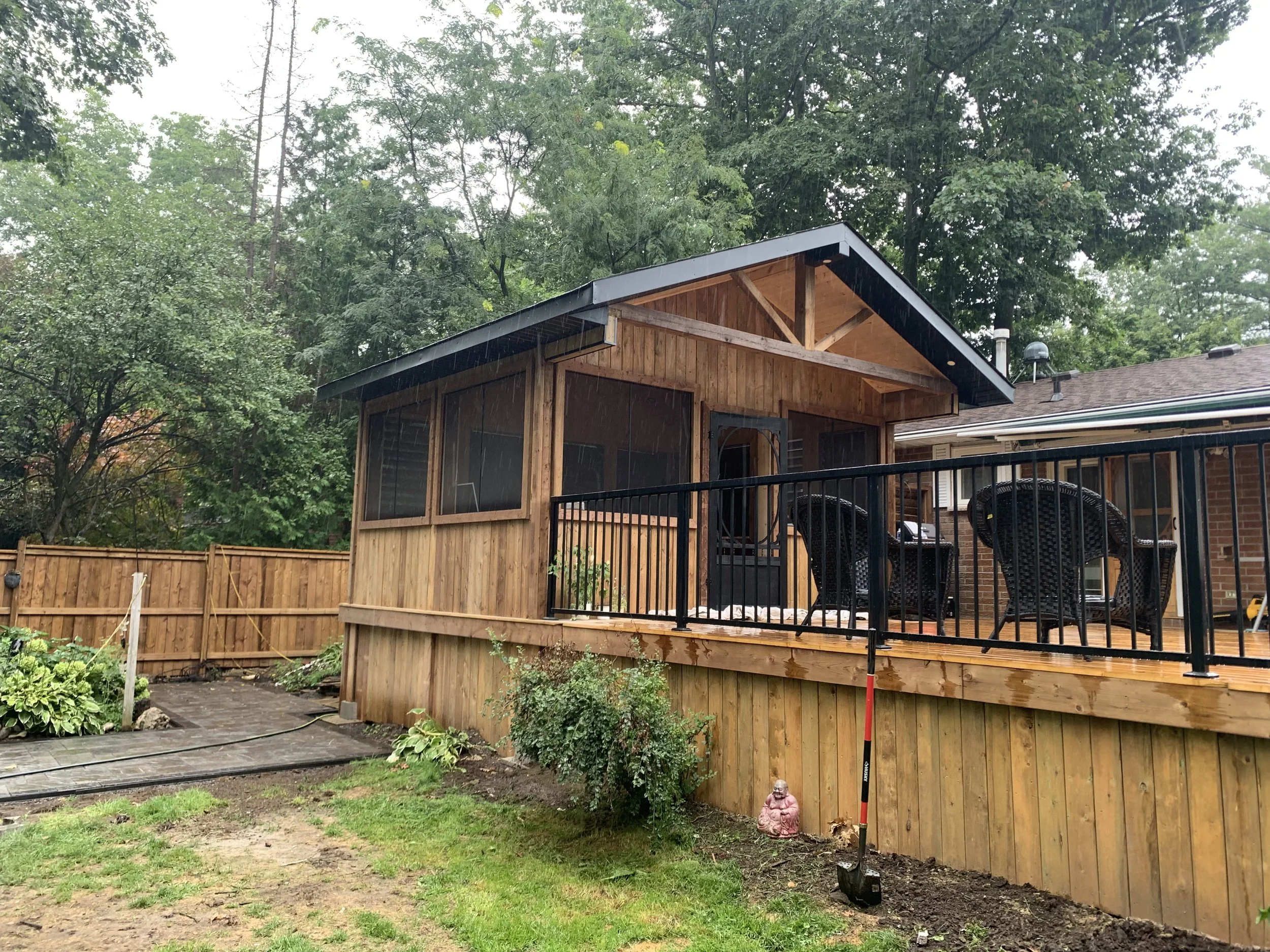 A backyard with a newly built wooden deck and a black metal railing, a small garden and a tree with green leaves; a wooden shed with a screened porch, a serving of chairs, and a Buddha sculpture in the yard; trees and overcast sky in the background.