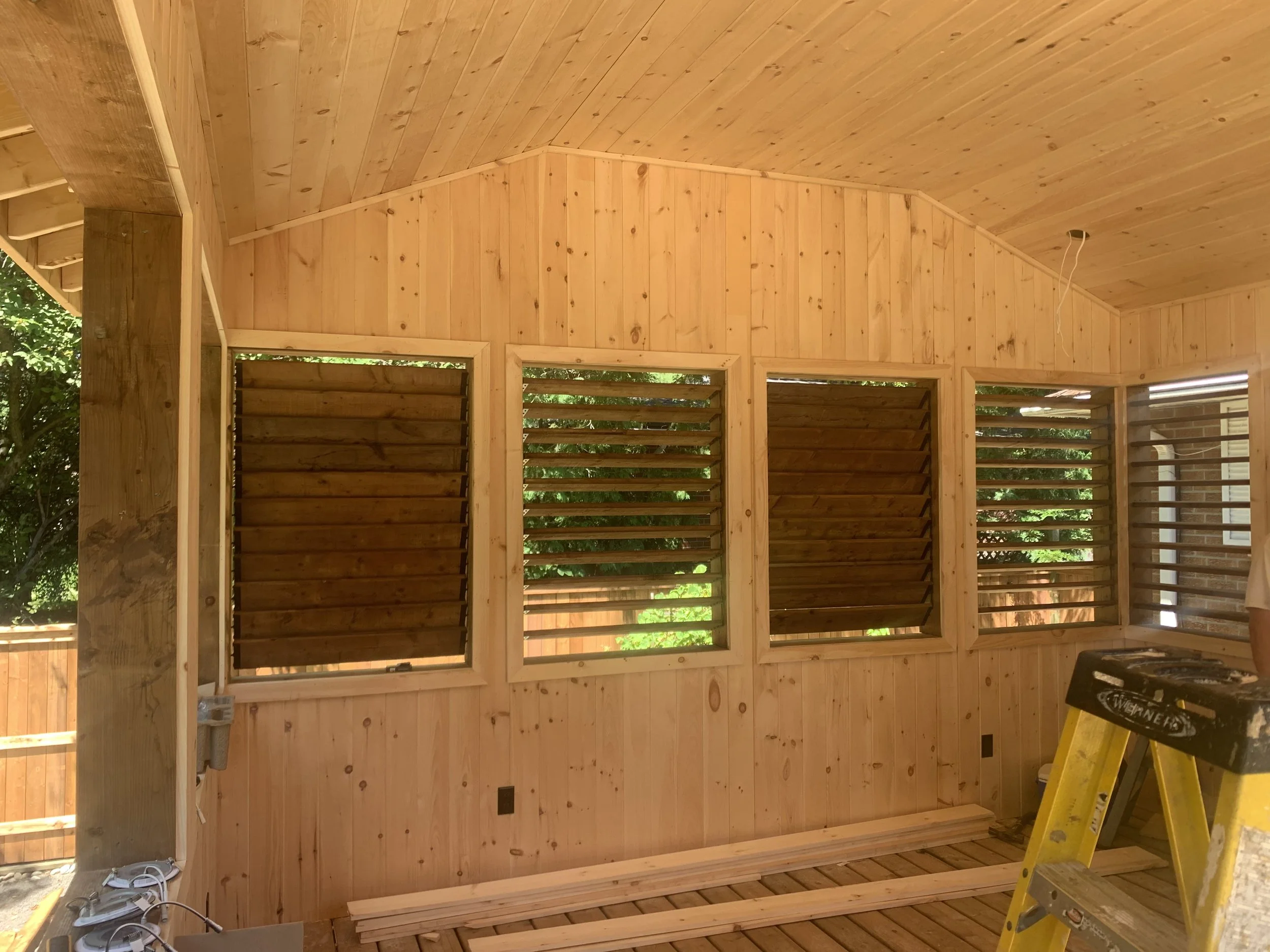 Interior of a wooden room or porch under construction, with unfinished wood paneling on walls and ceiling, construction tools and supplies visible.