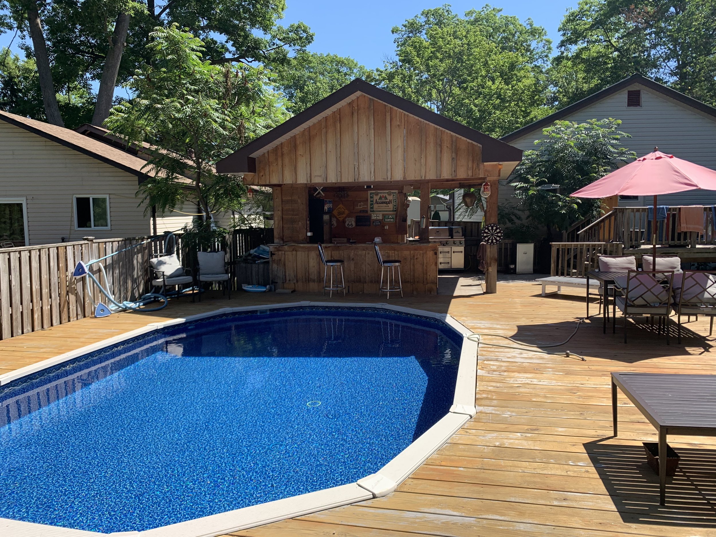 Backyard pool area with a wooden deck, pool, patio chairs, and a shaded bar with a grill, umbrella, and outdoor furniture, surrounded by trees and neighboring houses.
