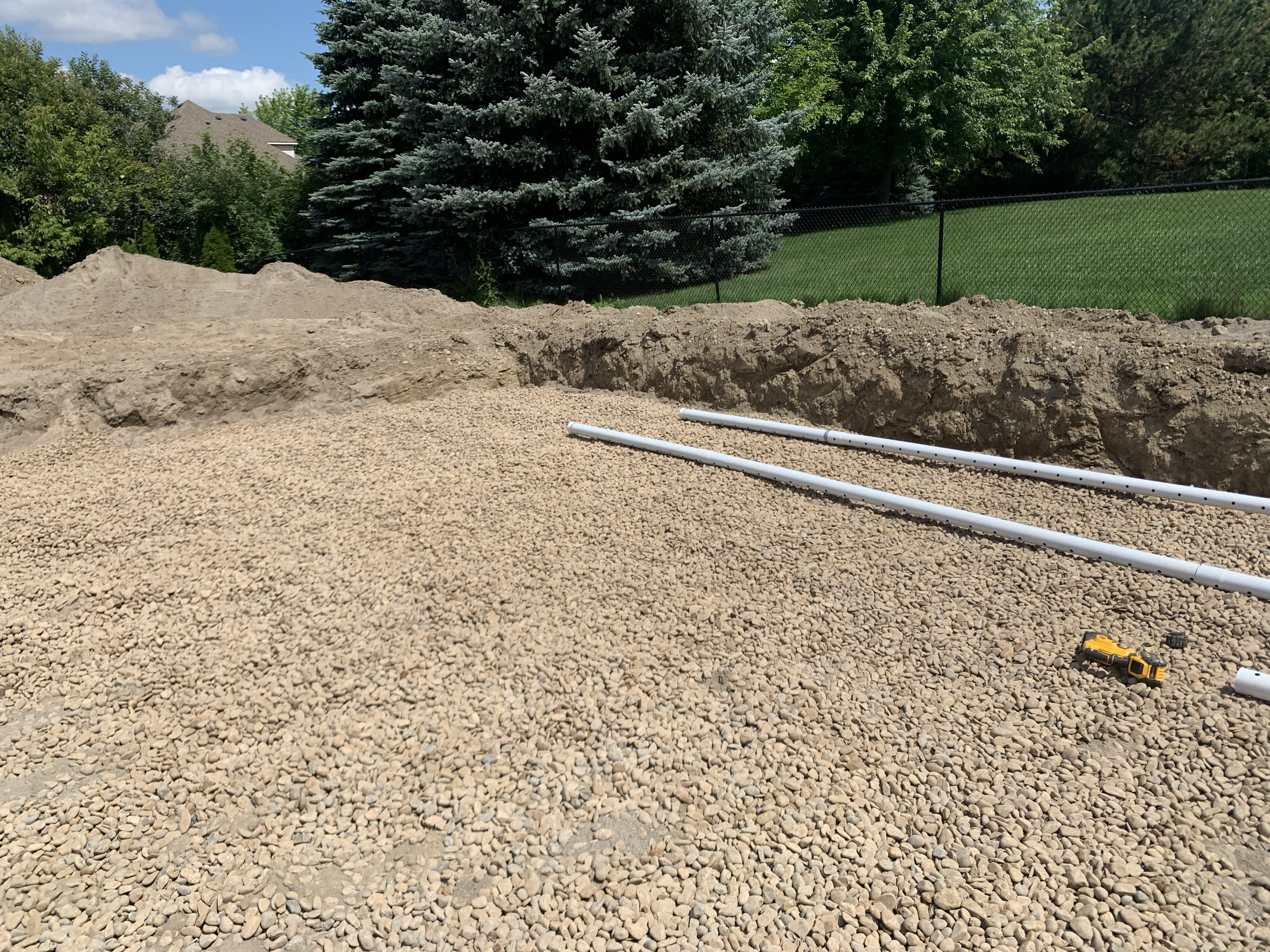 A backyard with a pile of dirt, black mesh fence, large evergreen tree, and green lawn. Construction tools and white PVC pipes are on a gravel surface in the foreground.