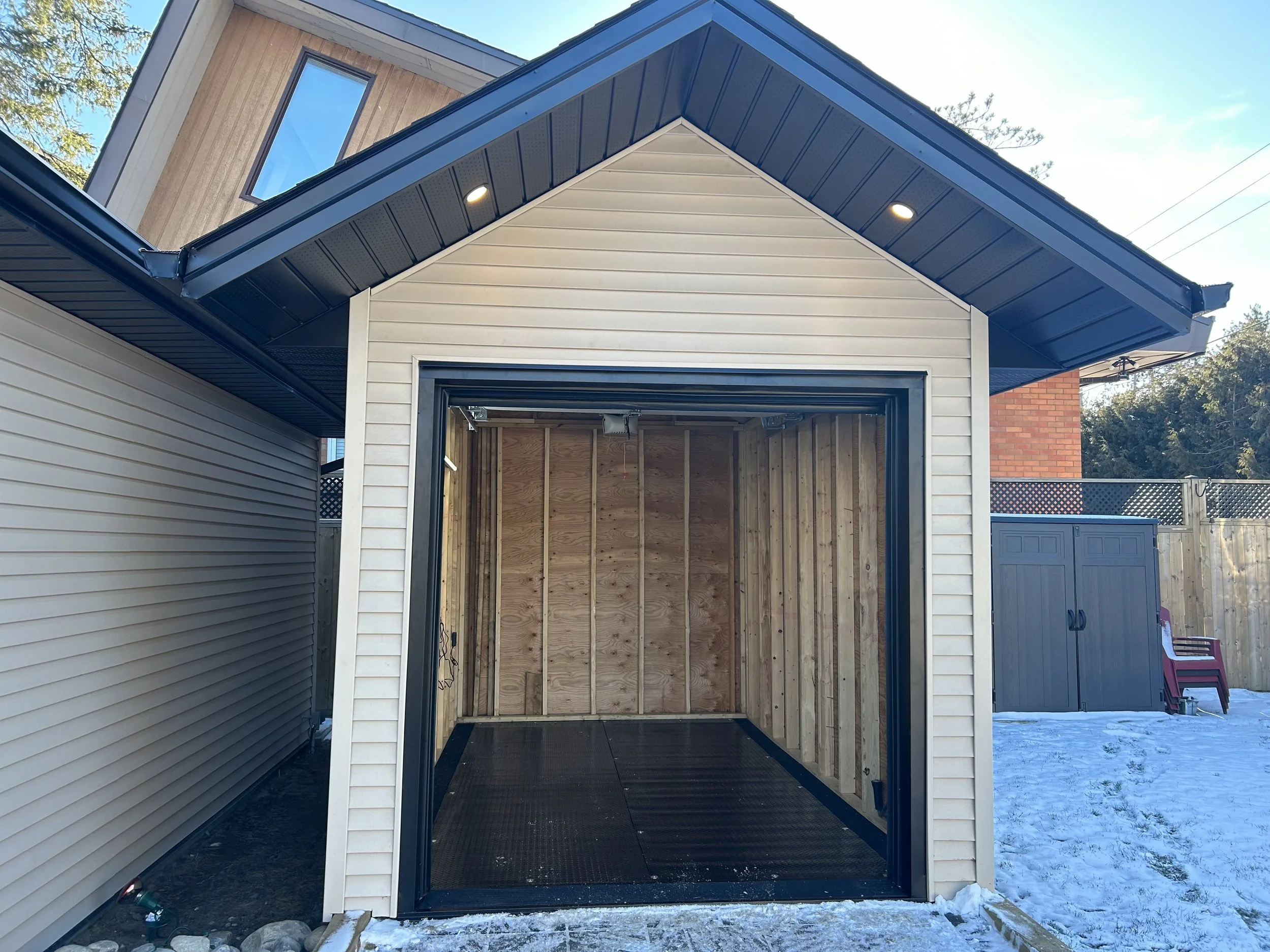 A garage with the front door open, showing an empty interior with wood framing. The garage has a black metal floor, white vinyl siding on the exterior, and a partially visible blue shed and snow on the ground outside.