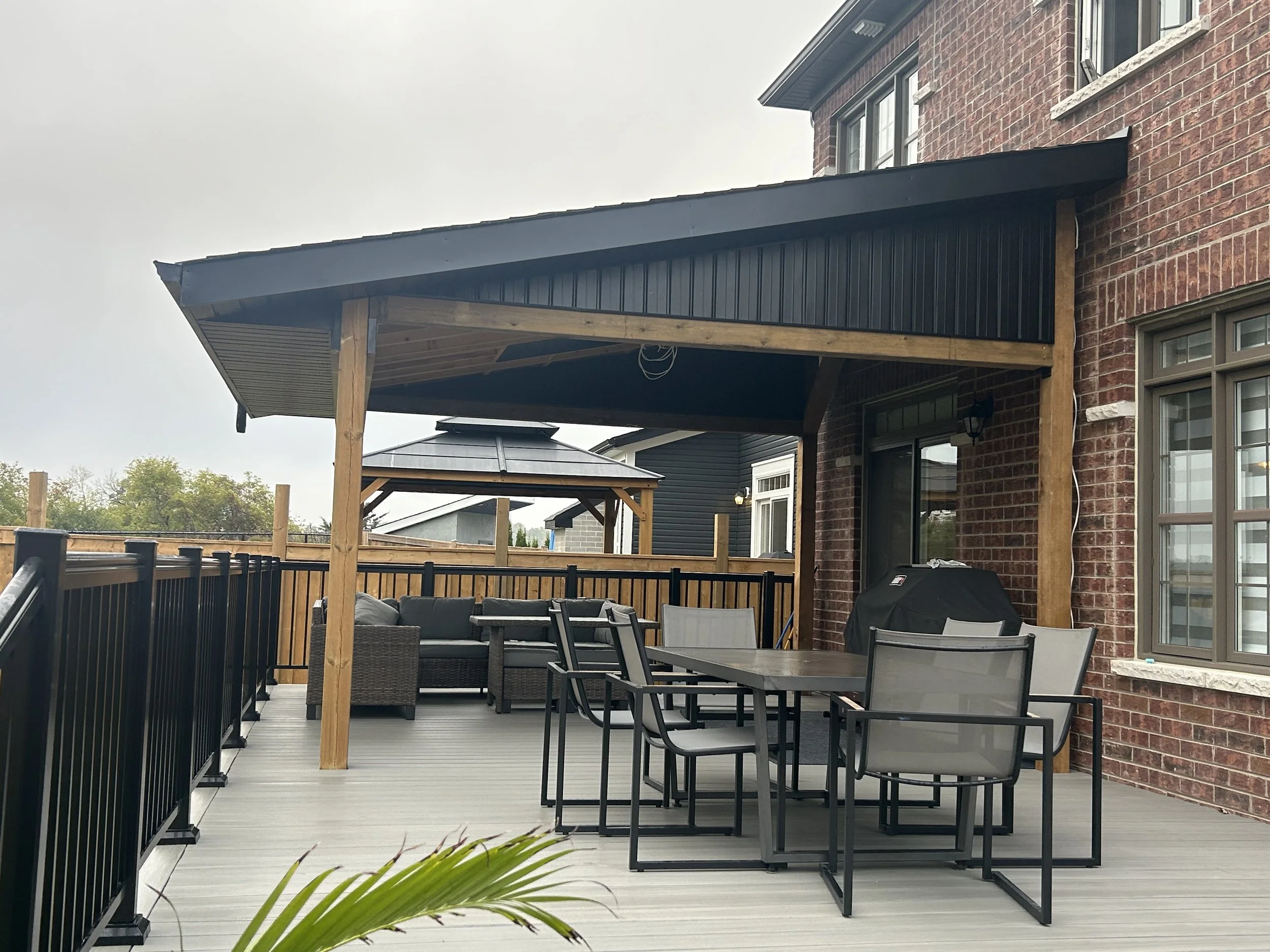A residential backyard patio with wooden and metal furniture, a privacy fence, a covered patio area with black ceiling, a brick house wall, and trees in the background.