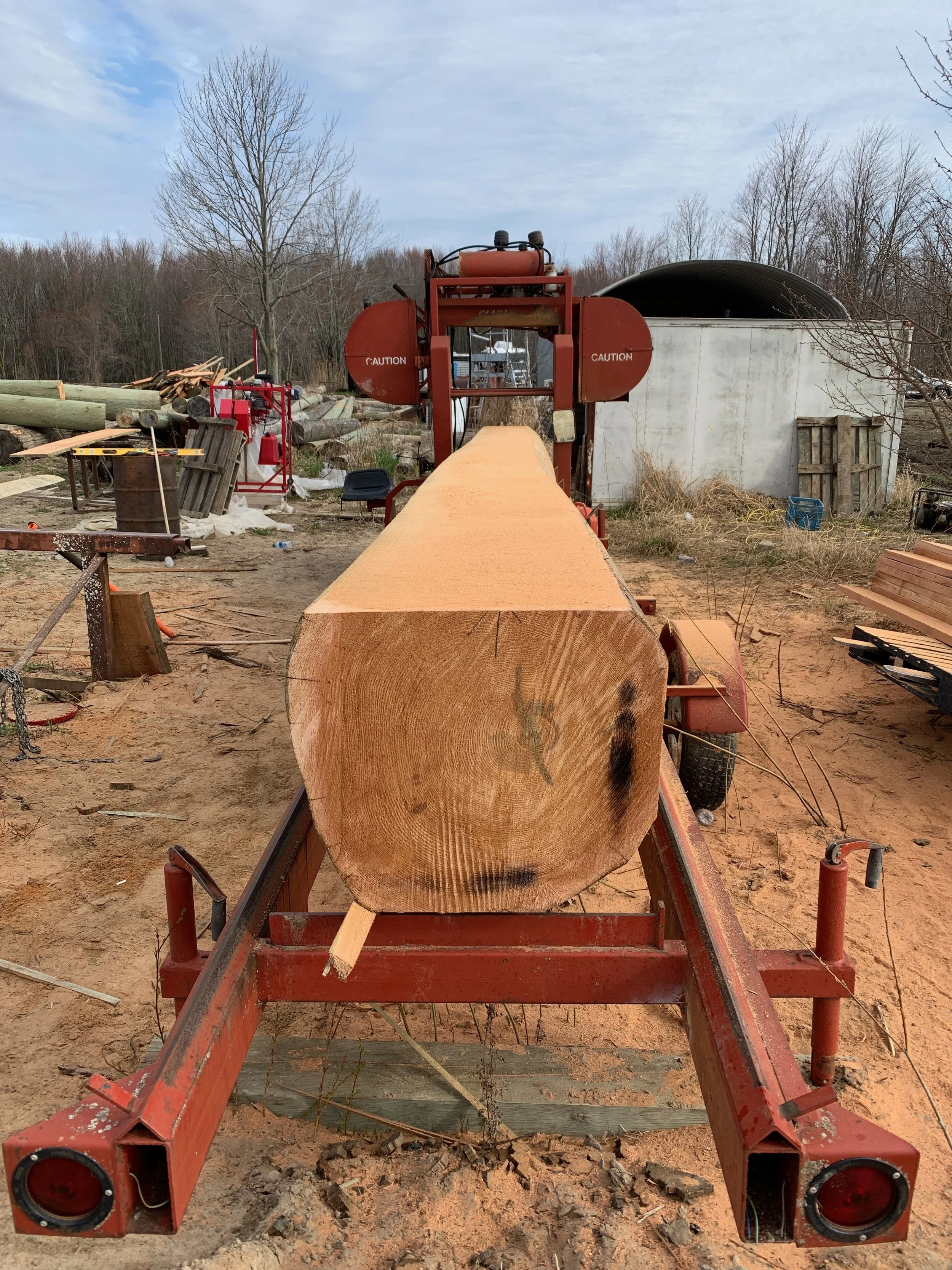 A large red lumber sawmill machine cutting a thick log into planks outdoors, with stacked logs and trees in the background, under a partly cloudy sky.
