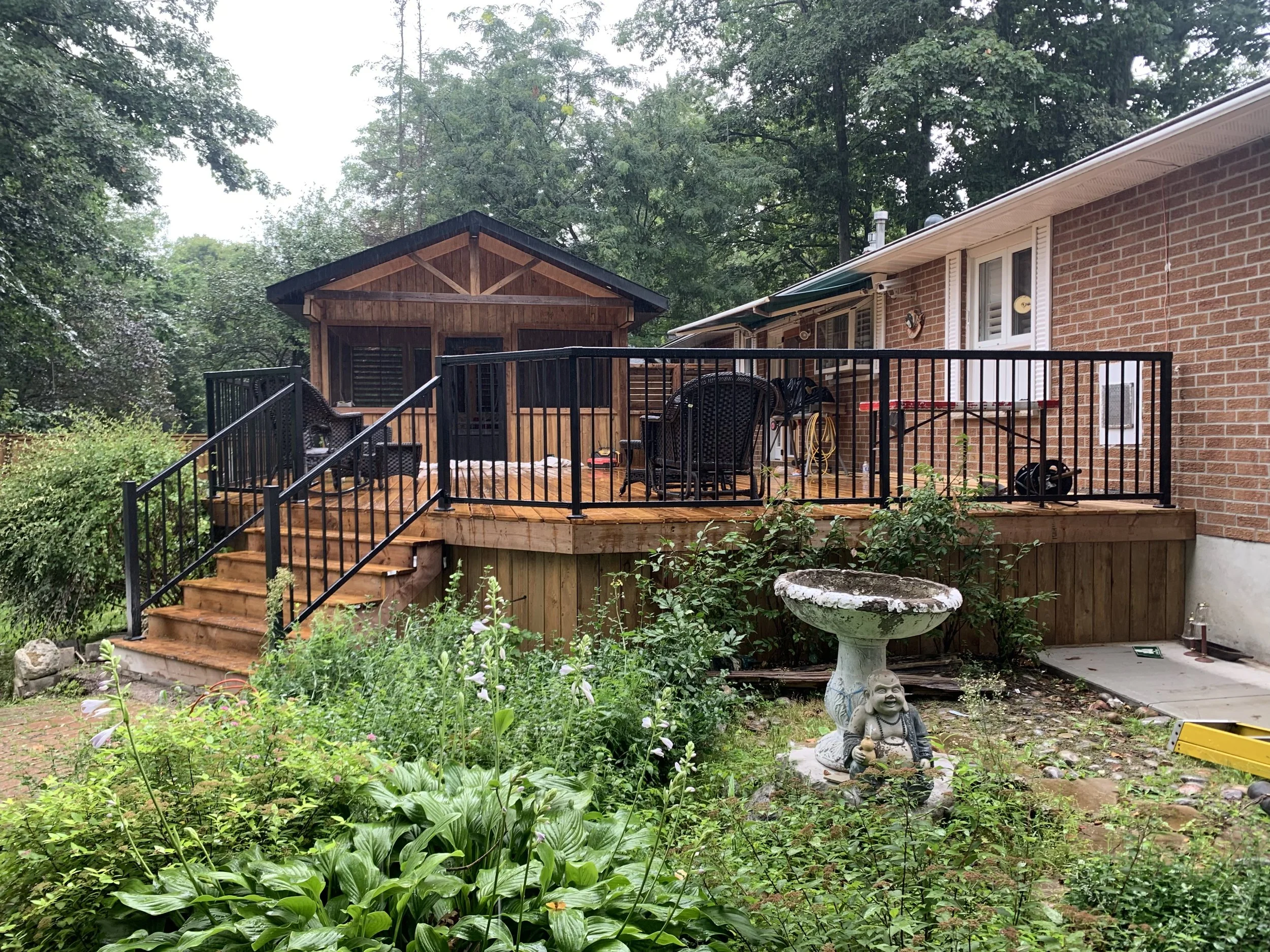 A backyard with a wooden deck, black metal railing, and outdoor furniture, adjacent to a brick house with a sloped roof. There are trees and greenery surrounding the area, along with a decorative garden statue of a smiling figure and statues, plants,