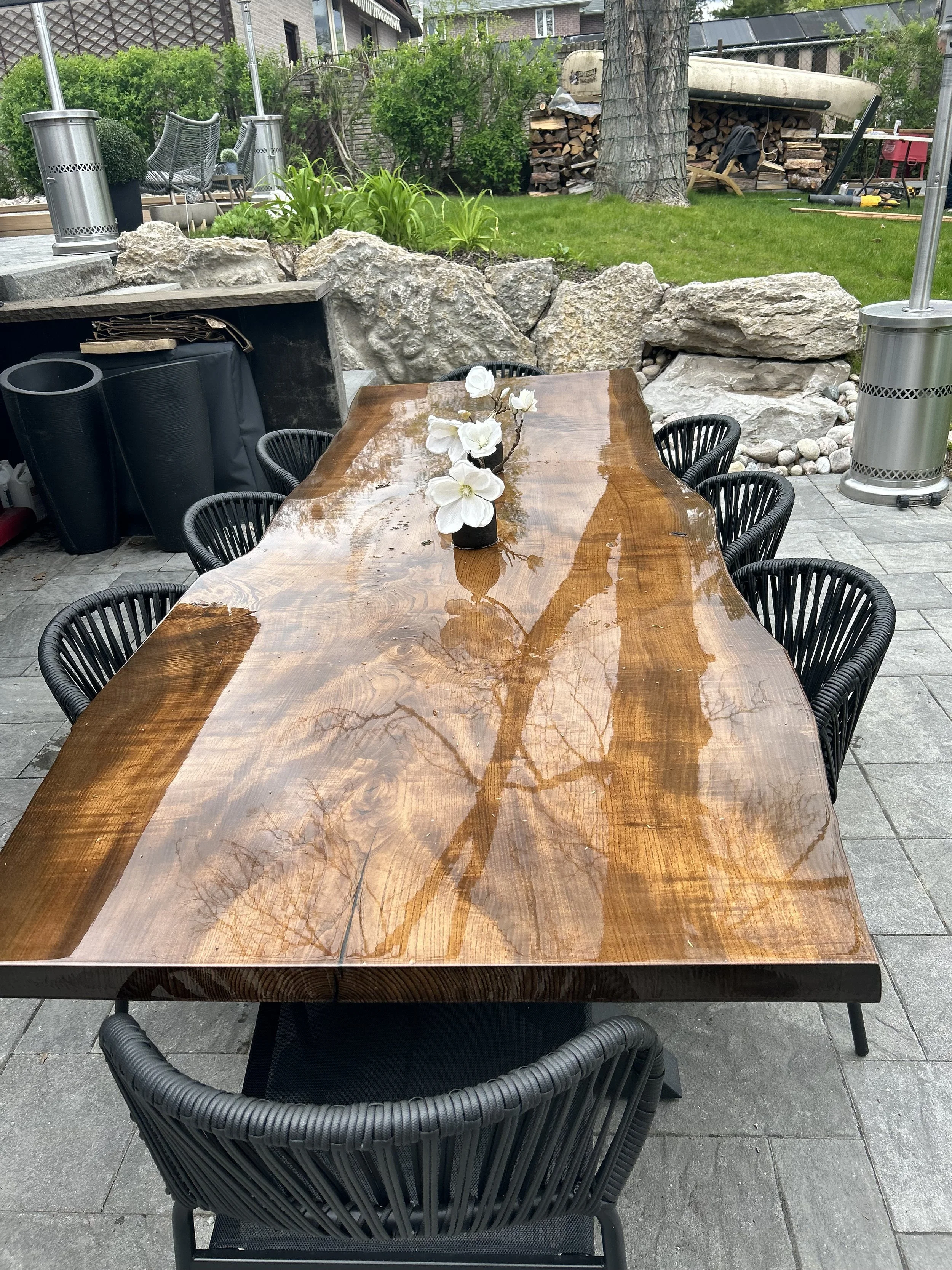Outdoor patio table with wet surface and flower centerpiece, surrounded by black woven chairs, in a backyard with green grass, trees, rocks, and gardening equipment.