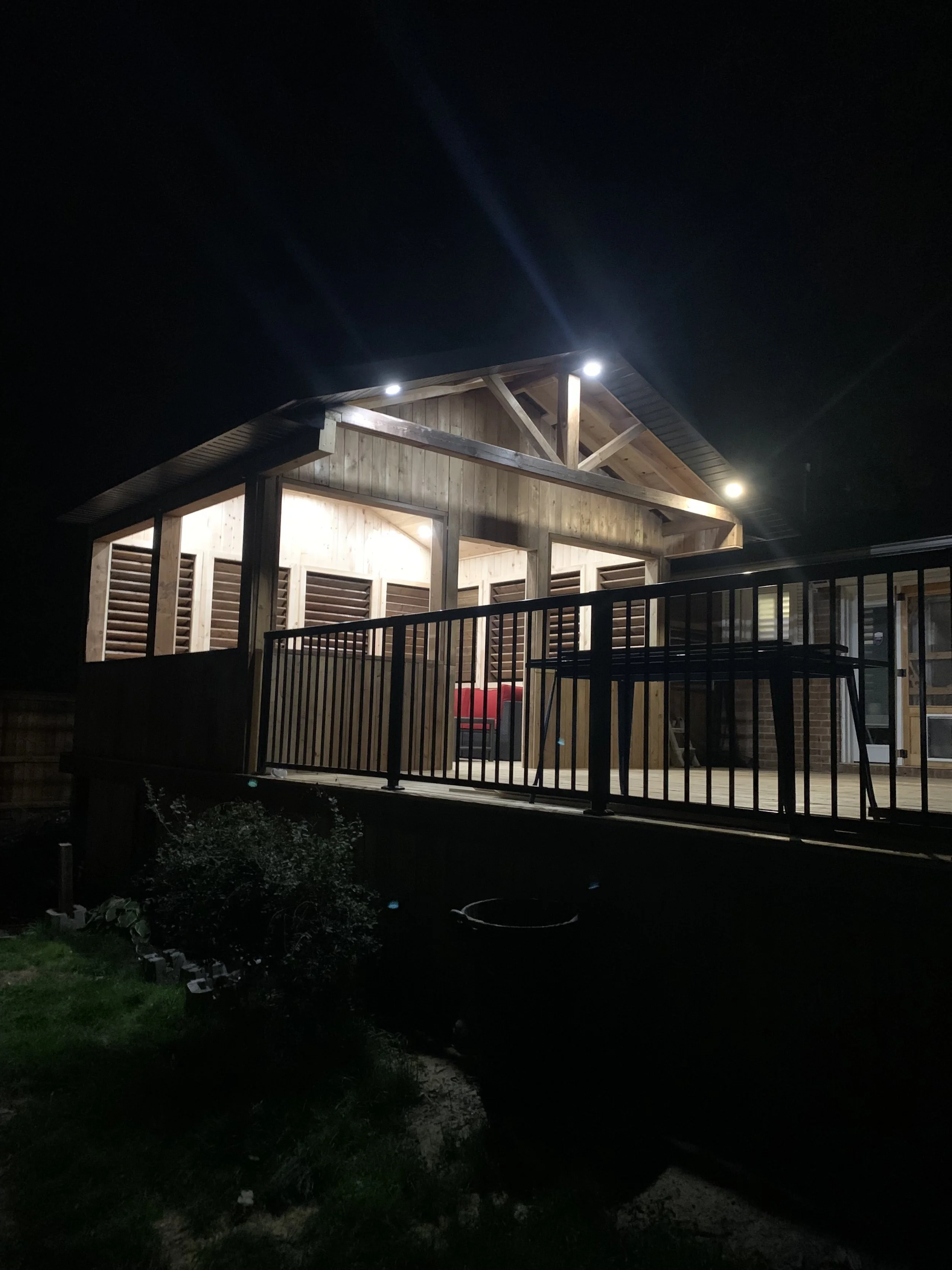 A wooden house patio at night with bright lights on the ceiling, a chair, and a black metal railing in the foreground. The surroundings are dark.