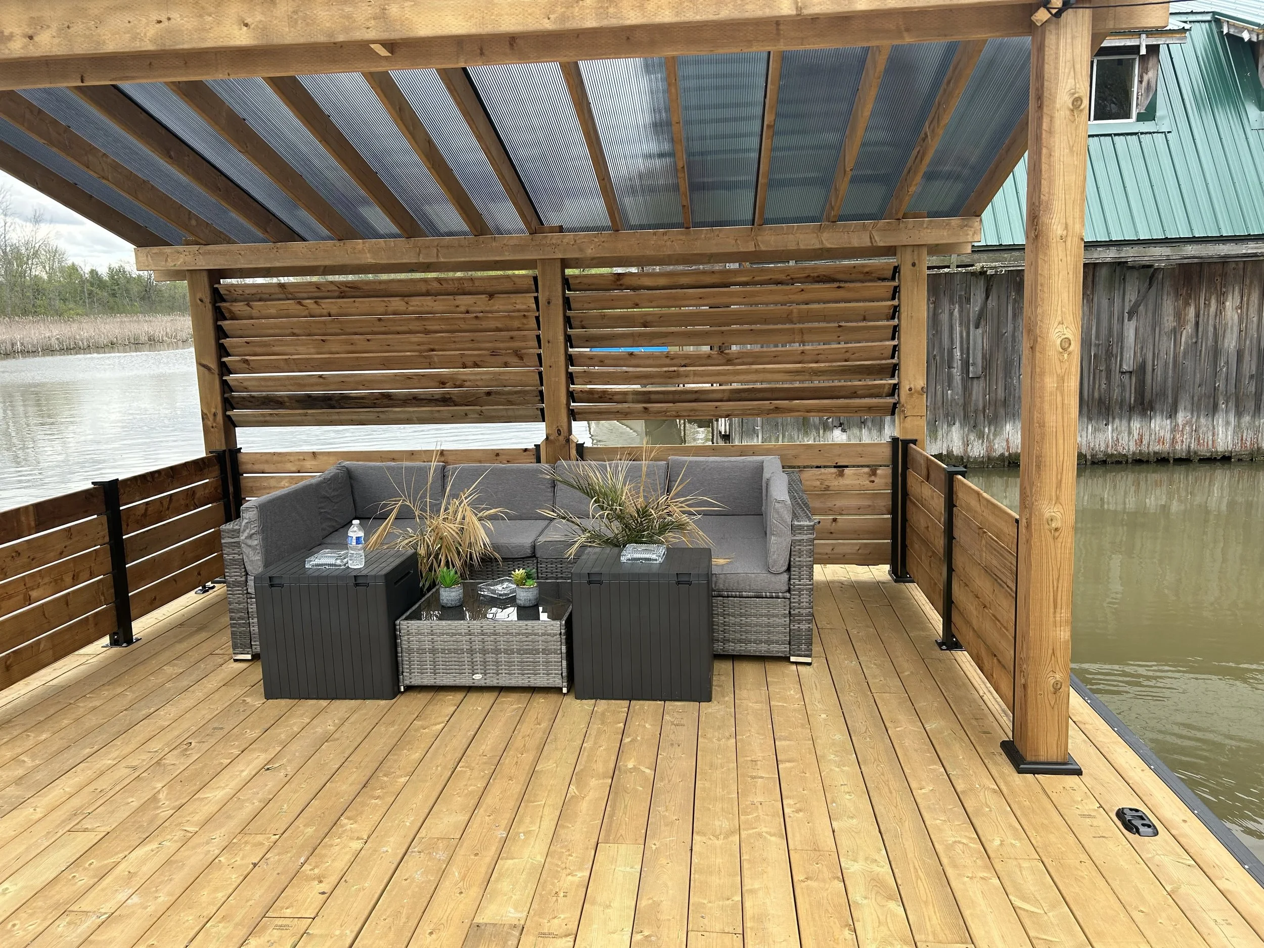 A wooden deck with a covered patio area overlooking a body of water with reeds in the background. There is outdoor furniture with cushions, plants, and decorative items on a black table.