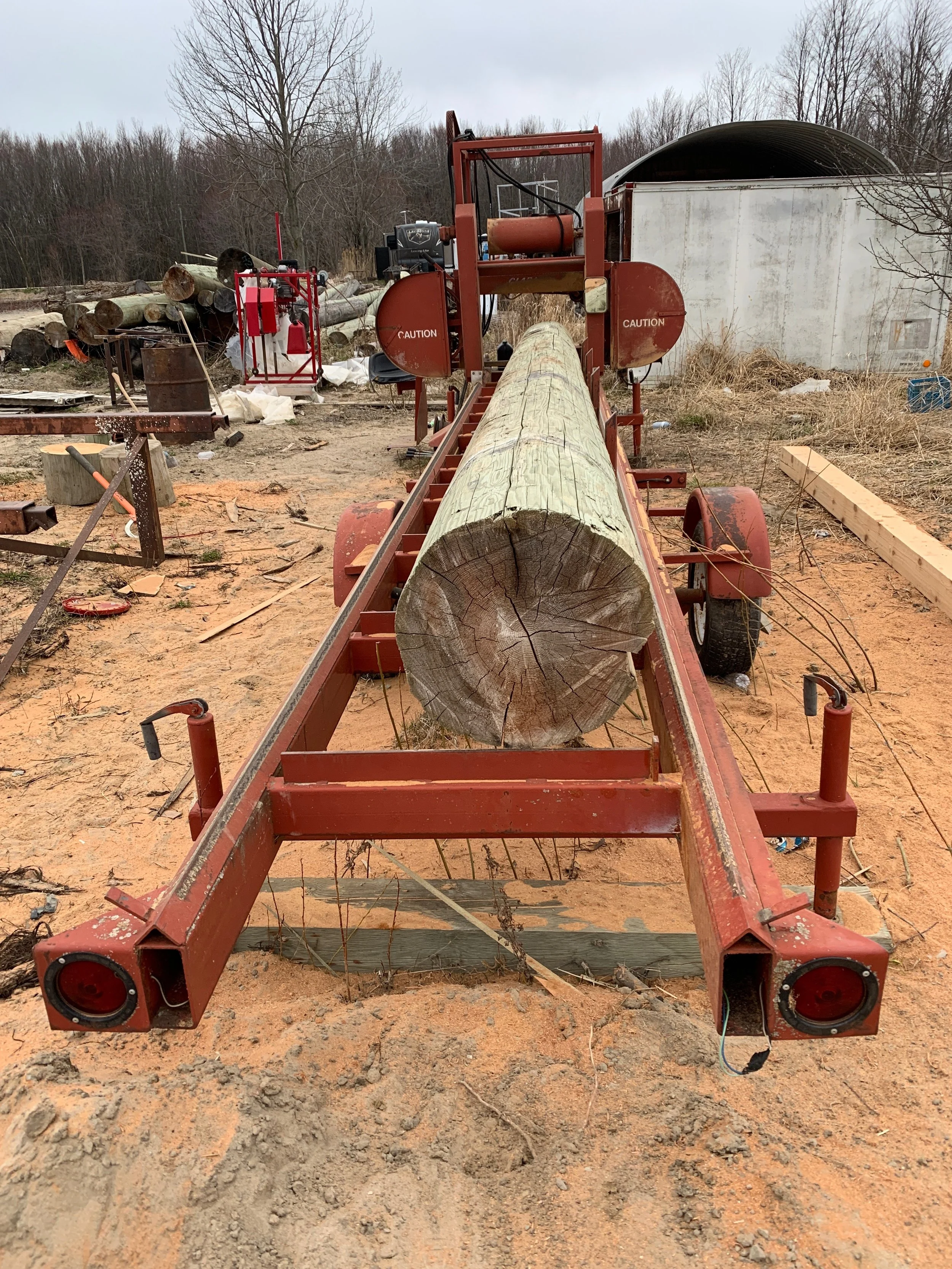 A large red and metal log splitter with a wood log loaded onto it, situated outdoors on a dirt ground, with trees and a storage shed in the background.