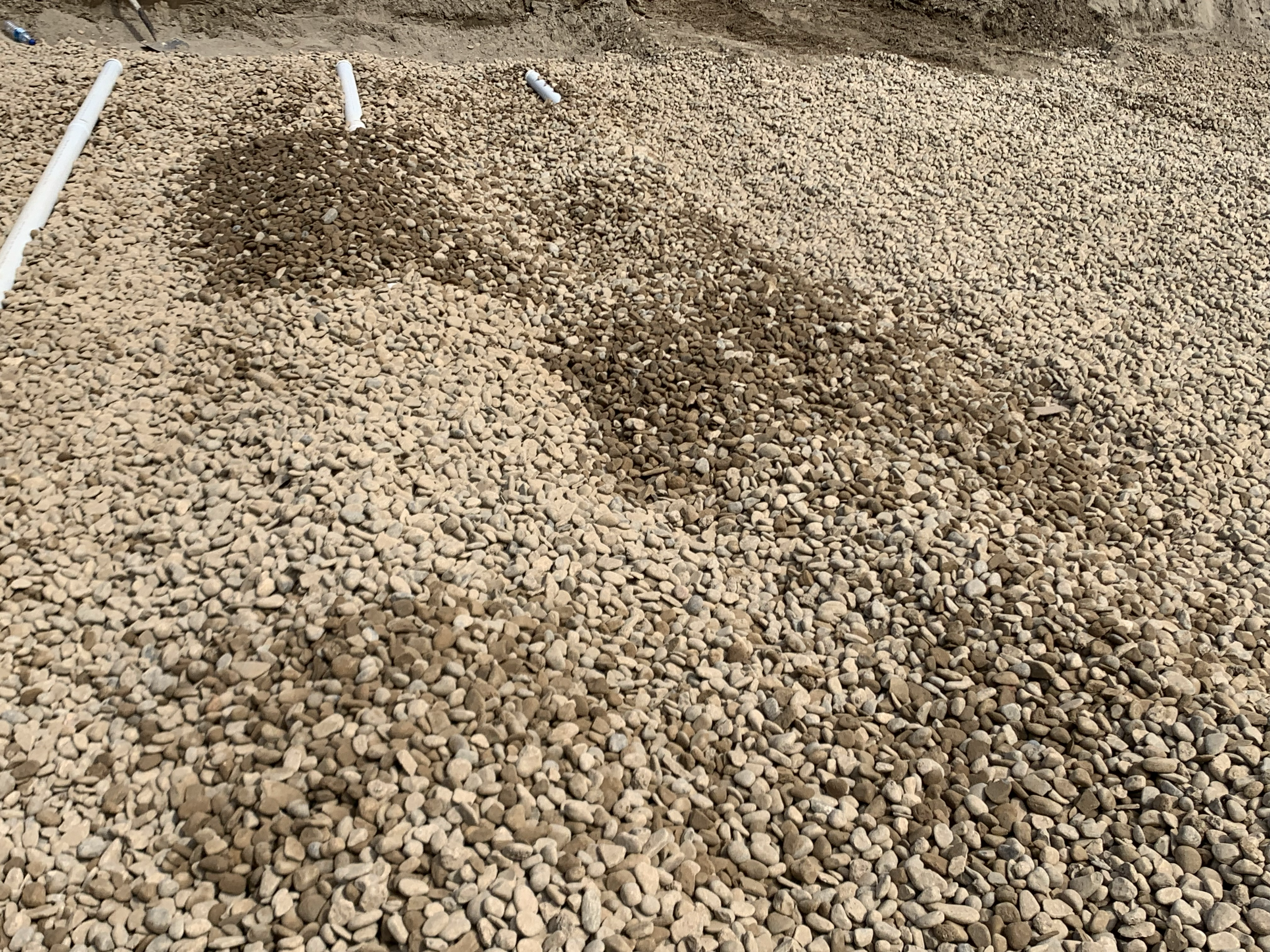 A pile of small, tan and gray gravel stones on a construction site or outdoor area with white pvc pipes on the left side.